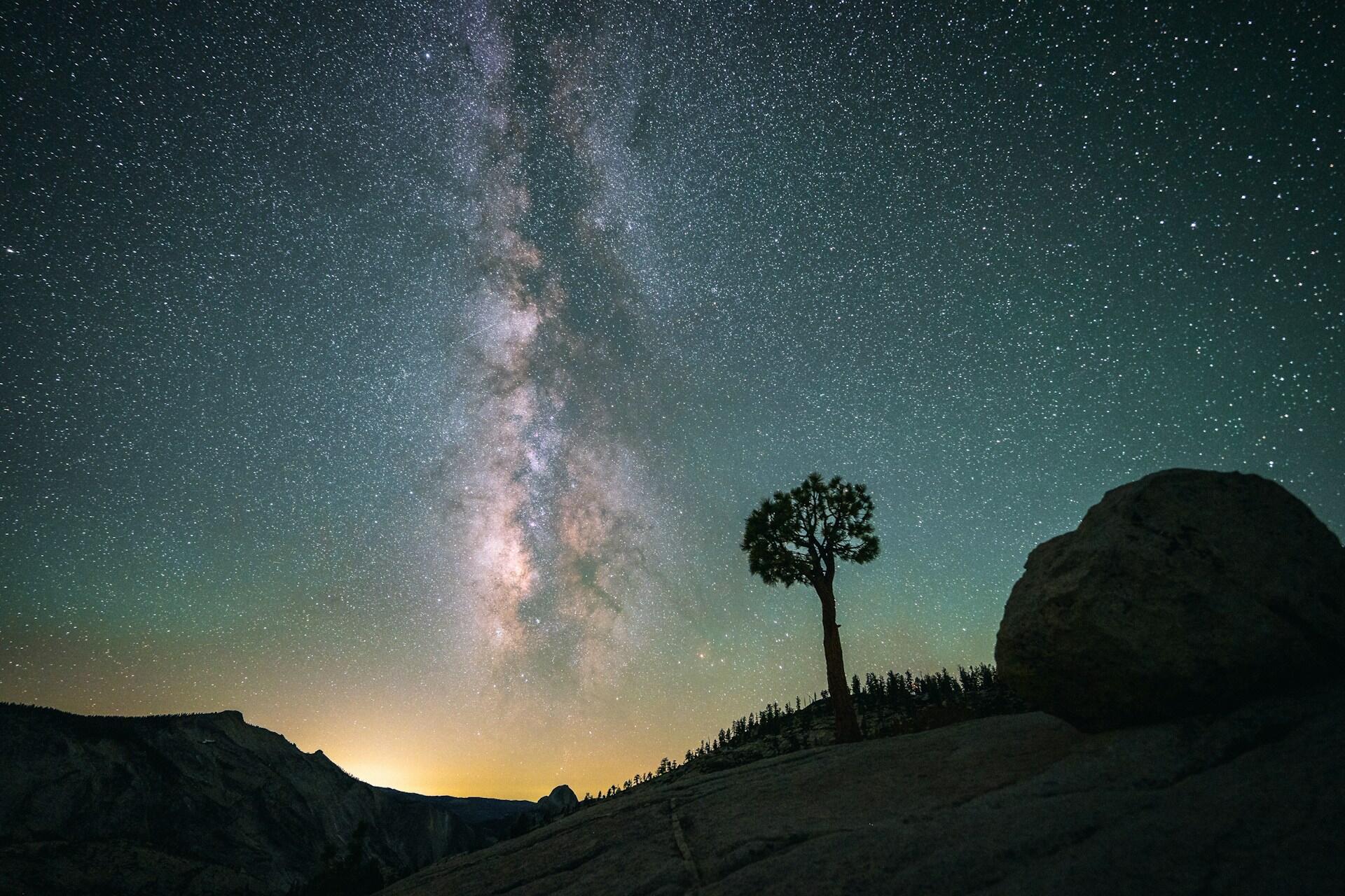 Galaxia de la Vía Láctea sobre un árbol solitario y una roca en un horizonte montañoso por la noche.