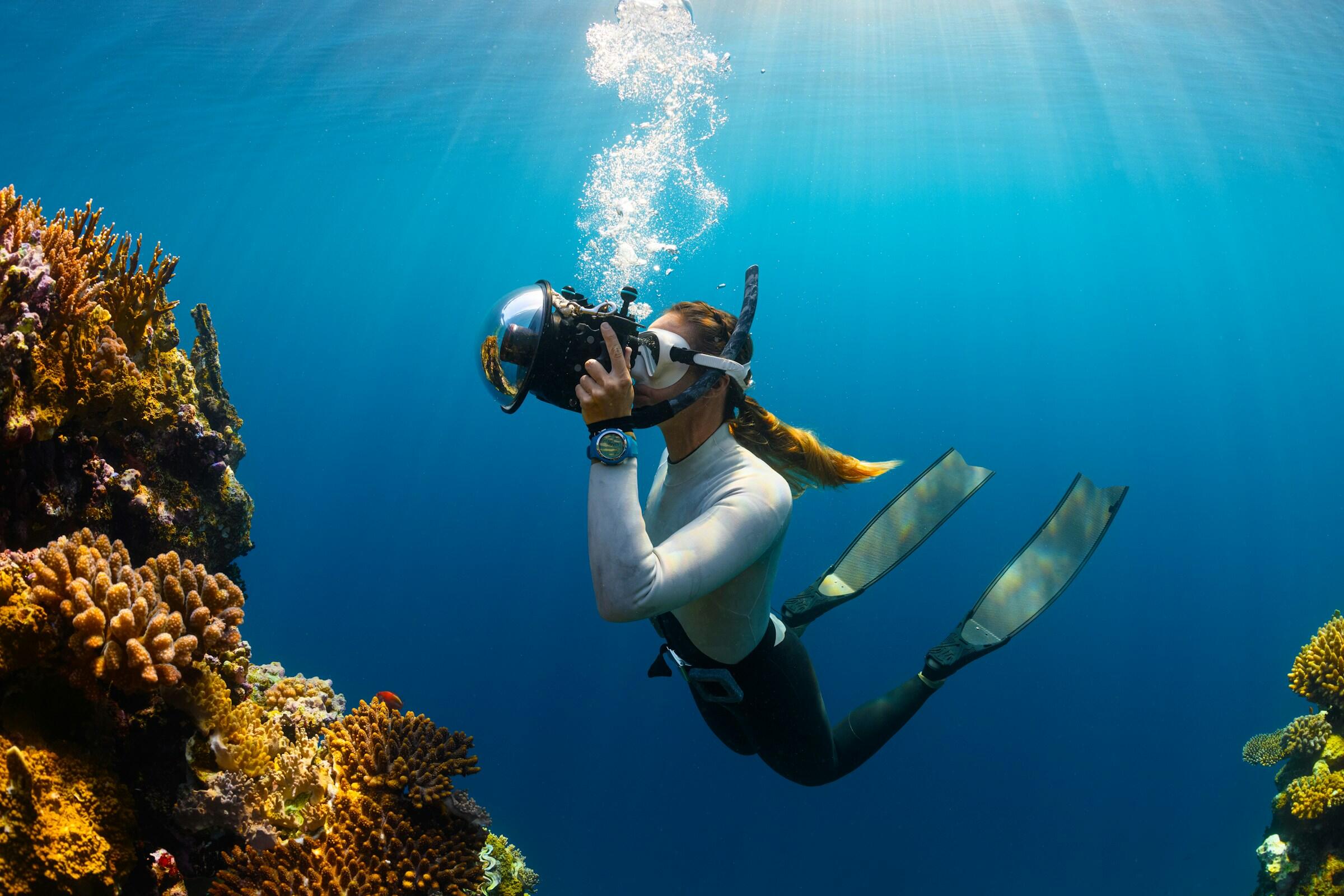 Un buceador bajo el agua con una carcasa de cámara tipo cúpula, rodeado de arrecifes de coral y bañado por la luz del sol que se filtra a través de las aguas azules y cristalinas.