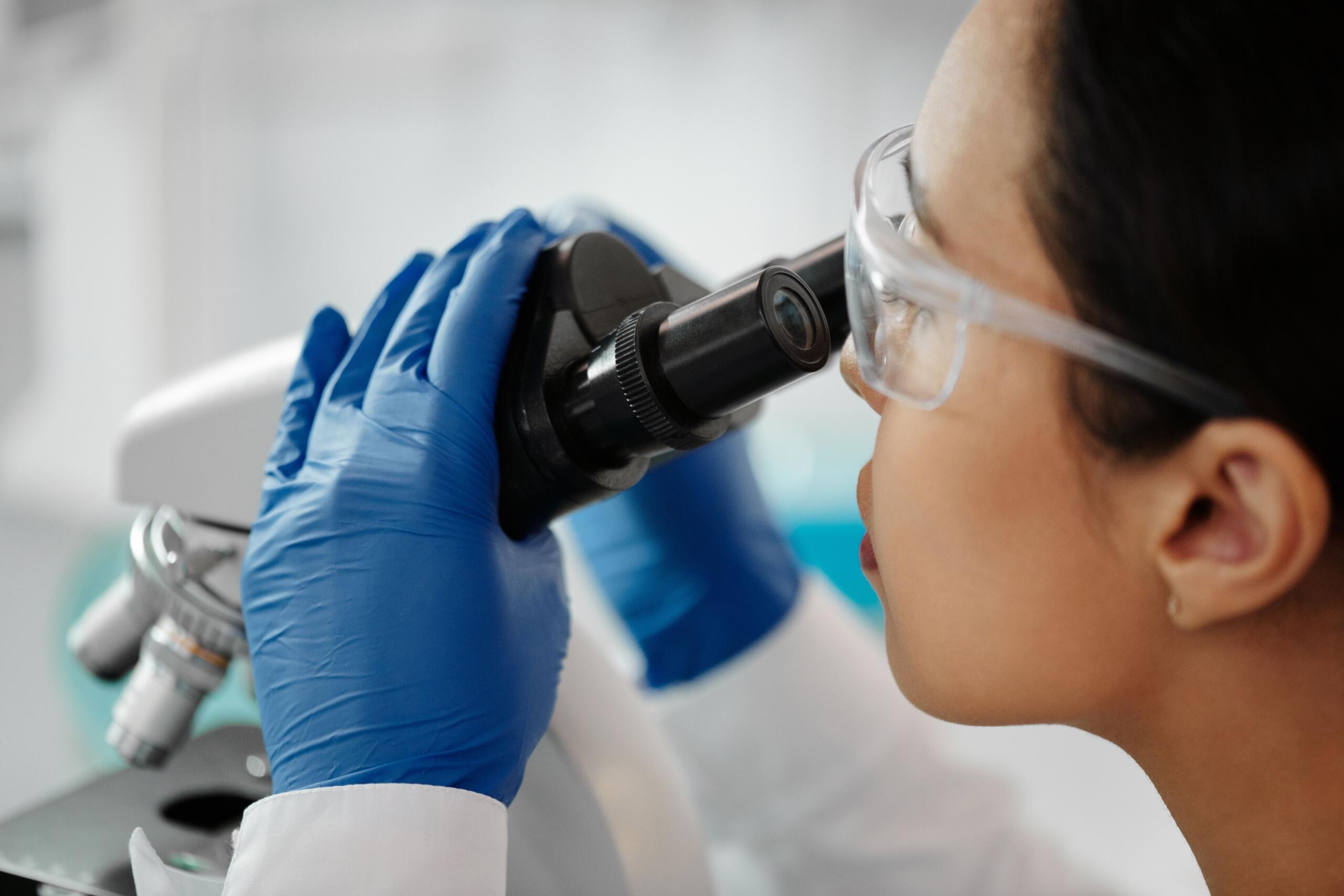 Primer plano de una científica joven y concentrada, equipada con gafas de seguridad y guantes de nitrilo azules, observando atentamente a través de un microscopio en un entorno de laboratorio limpio.
