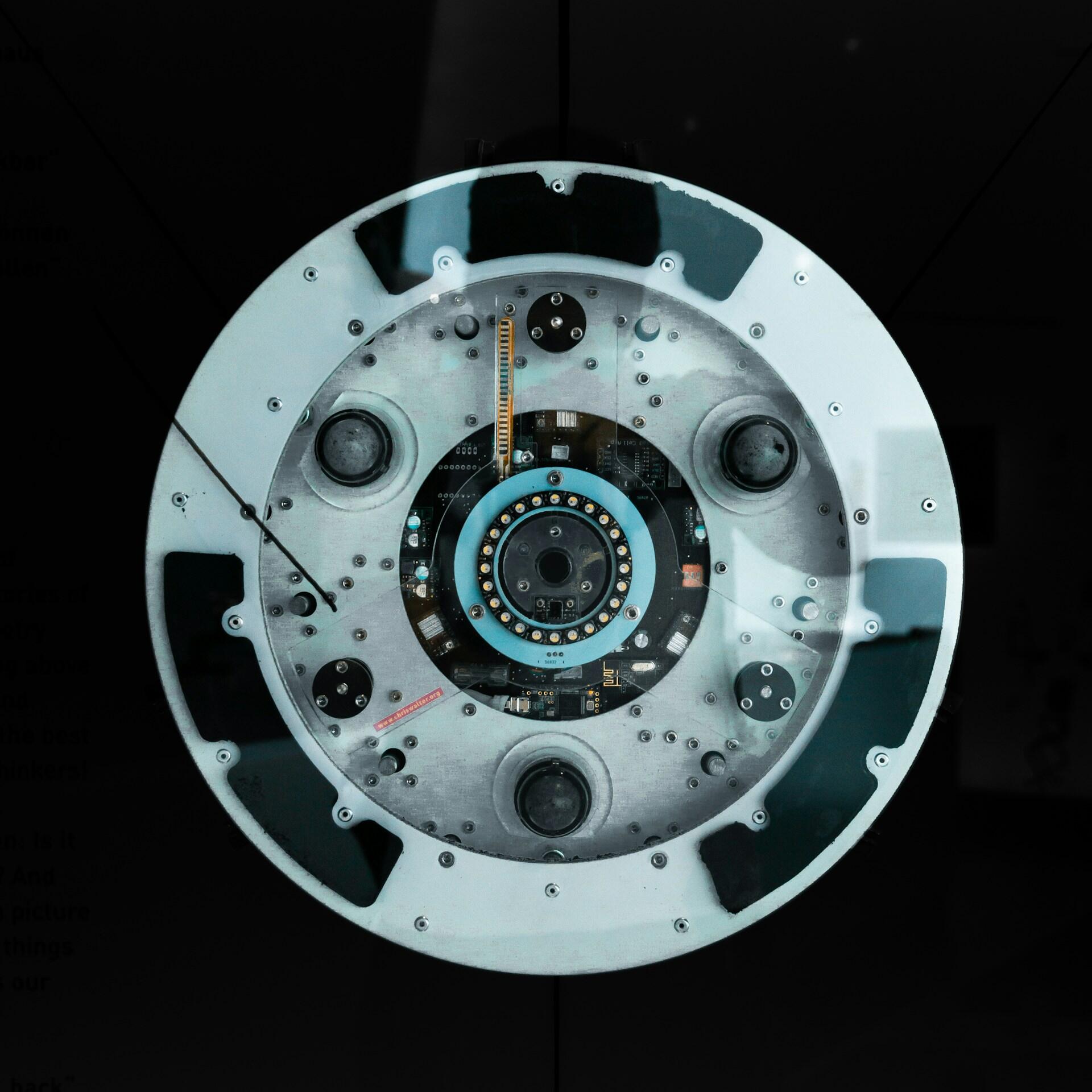 Vista desde arriba de un dispositivo mecánico circular con circuitos, tornillos y sensores a la vista, y detalles en azul sobre un fondo oscuro; tono técnico.