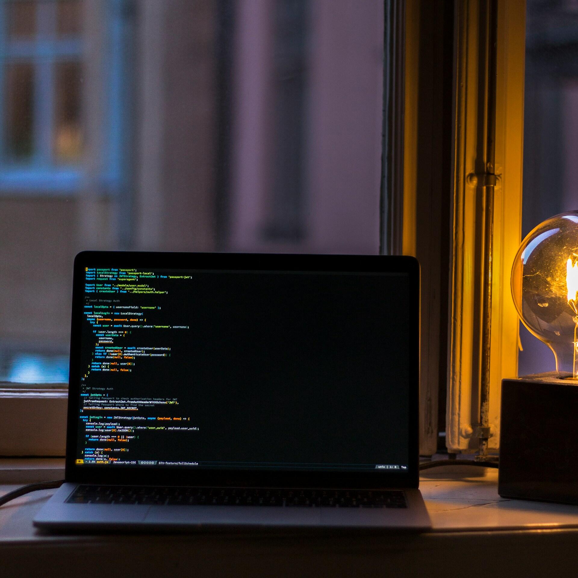 Un ordenador portátil en el que se ve un código de colores descansa junto a una ventana, al lado de una lámpara esférica que emite una luz cálida, creando un espacio de trabajo nocturno acogedor y evocador.