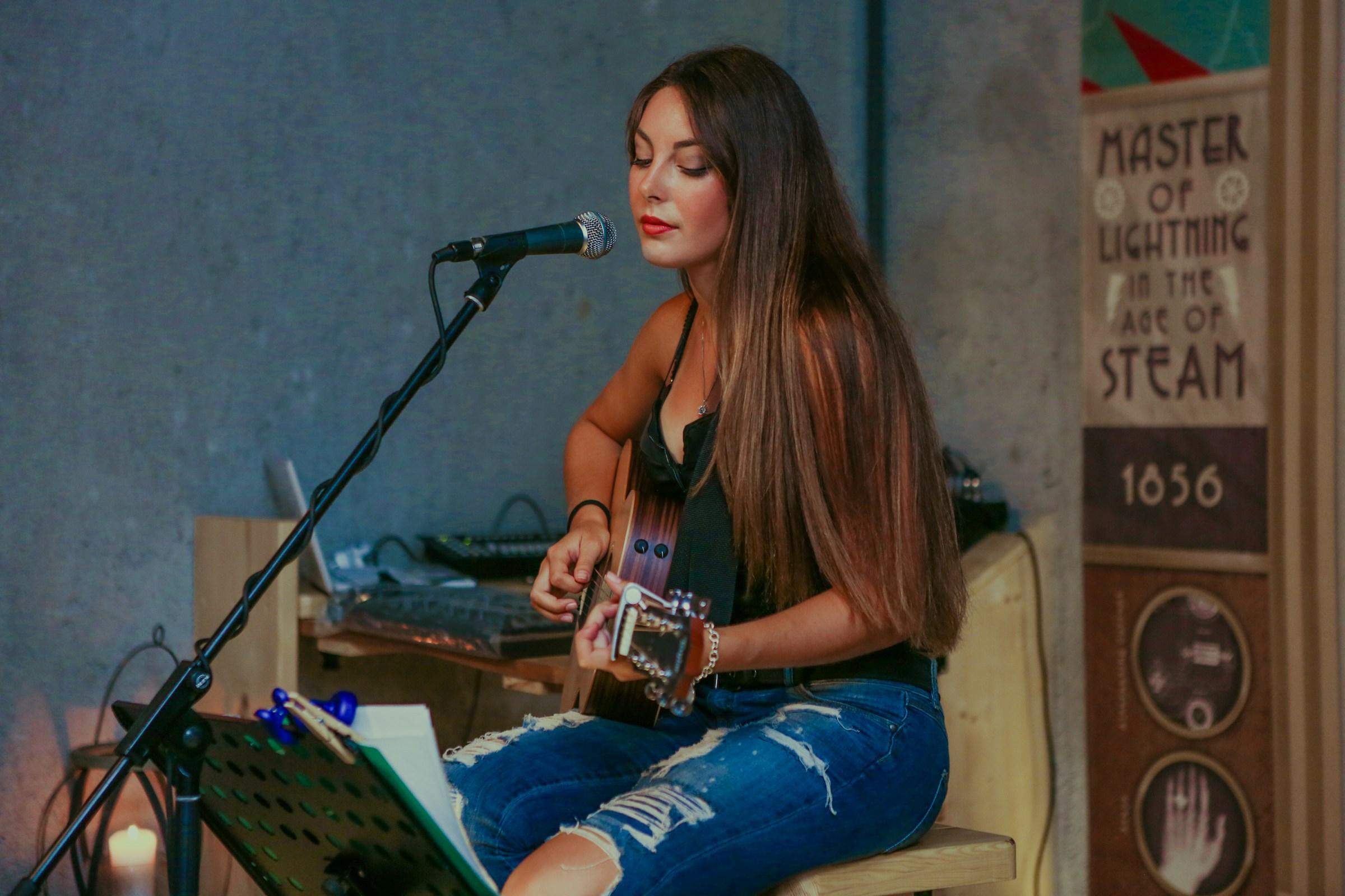 Mujer tocando la guitarra sentada junto a un micrófono y un atril.