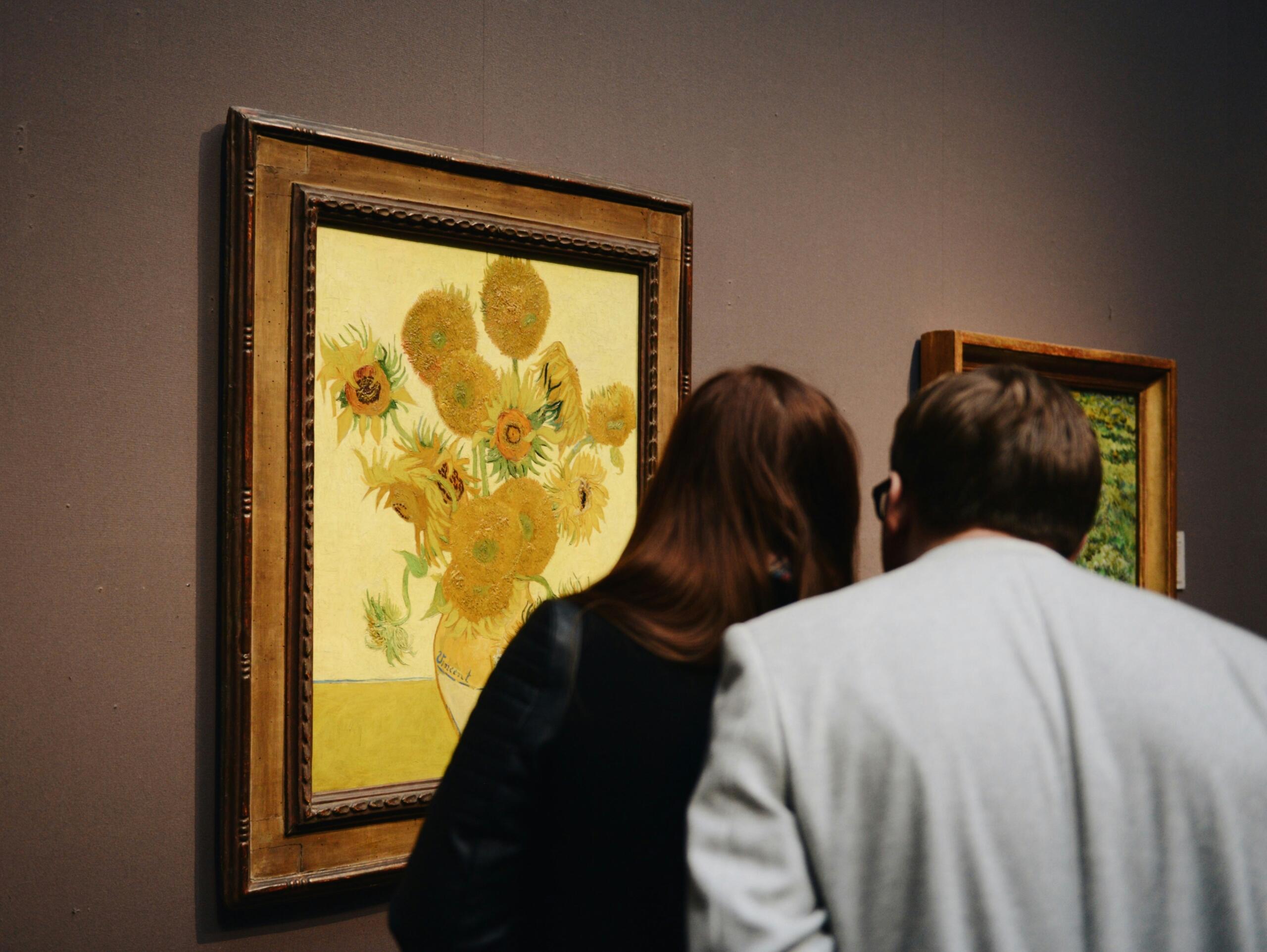 Una pareja admira el cuadro «Girasoles» de Van Gogh en una galería, con las cabezas muy juntas para apreciar la obra.