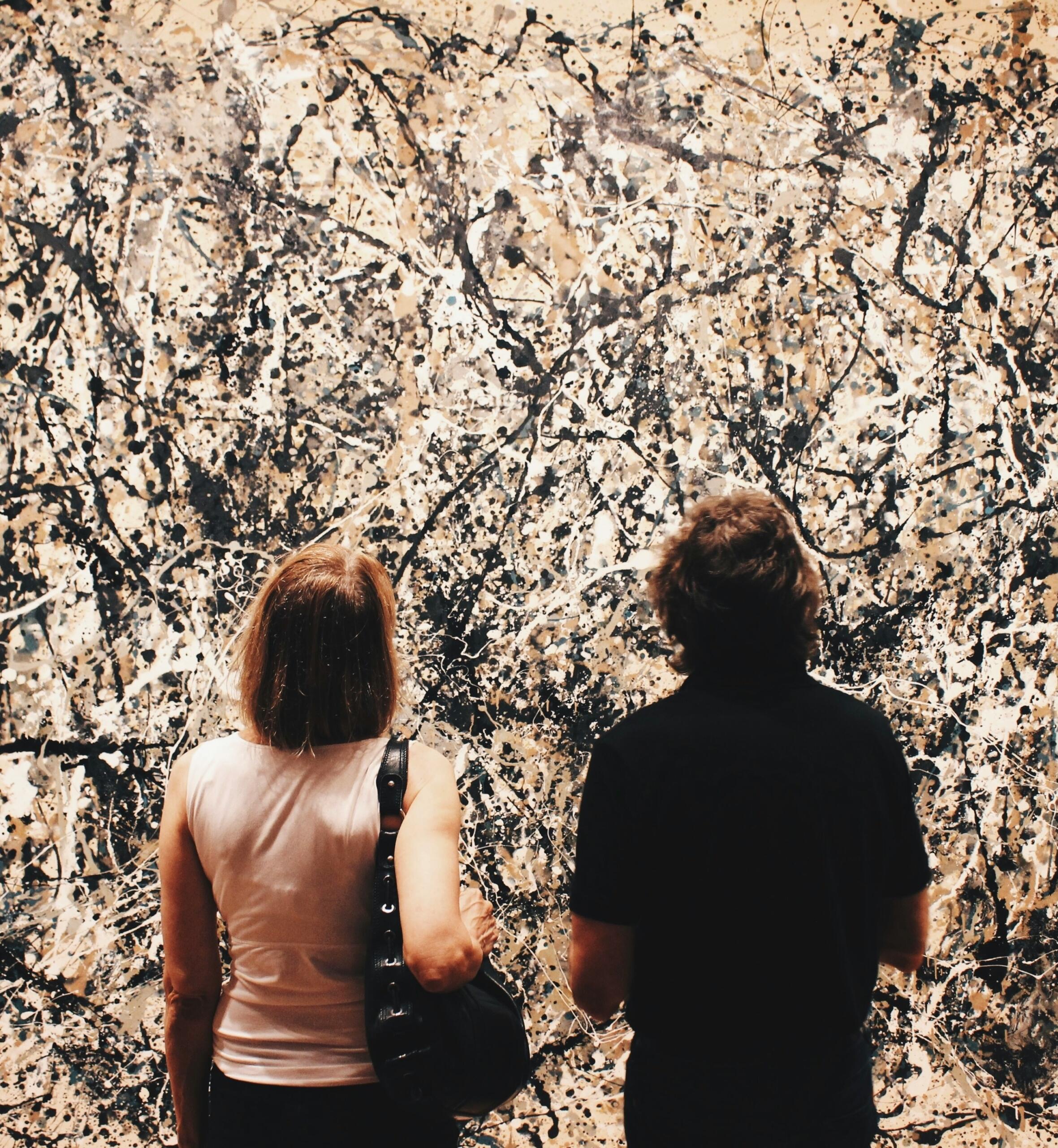 Dos visitantes del museo admiran un gran cuadro abstracto de jackson pollok  con caóticas salpicaduras en blanco y negro sobre un fondo beige.