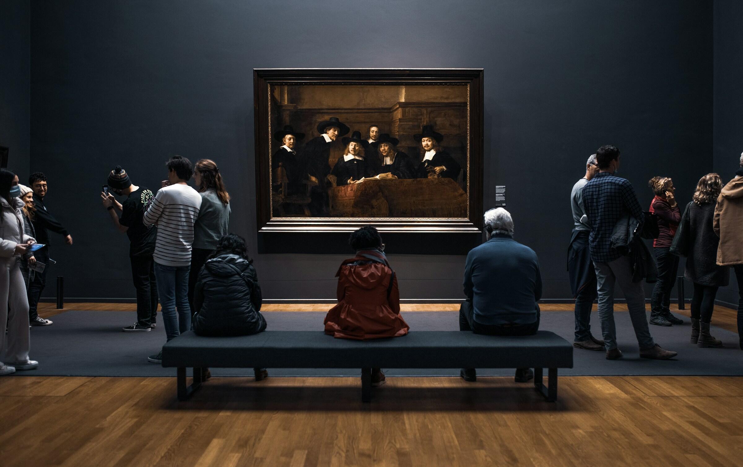 Los visitantes admiran un cuadro famoso de Rembrandt  en un museo poco iluminado, rodeado de paredes oscuras y cálidos suelos de madera.