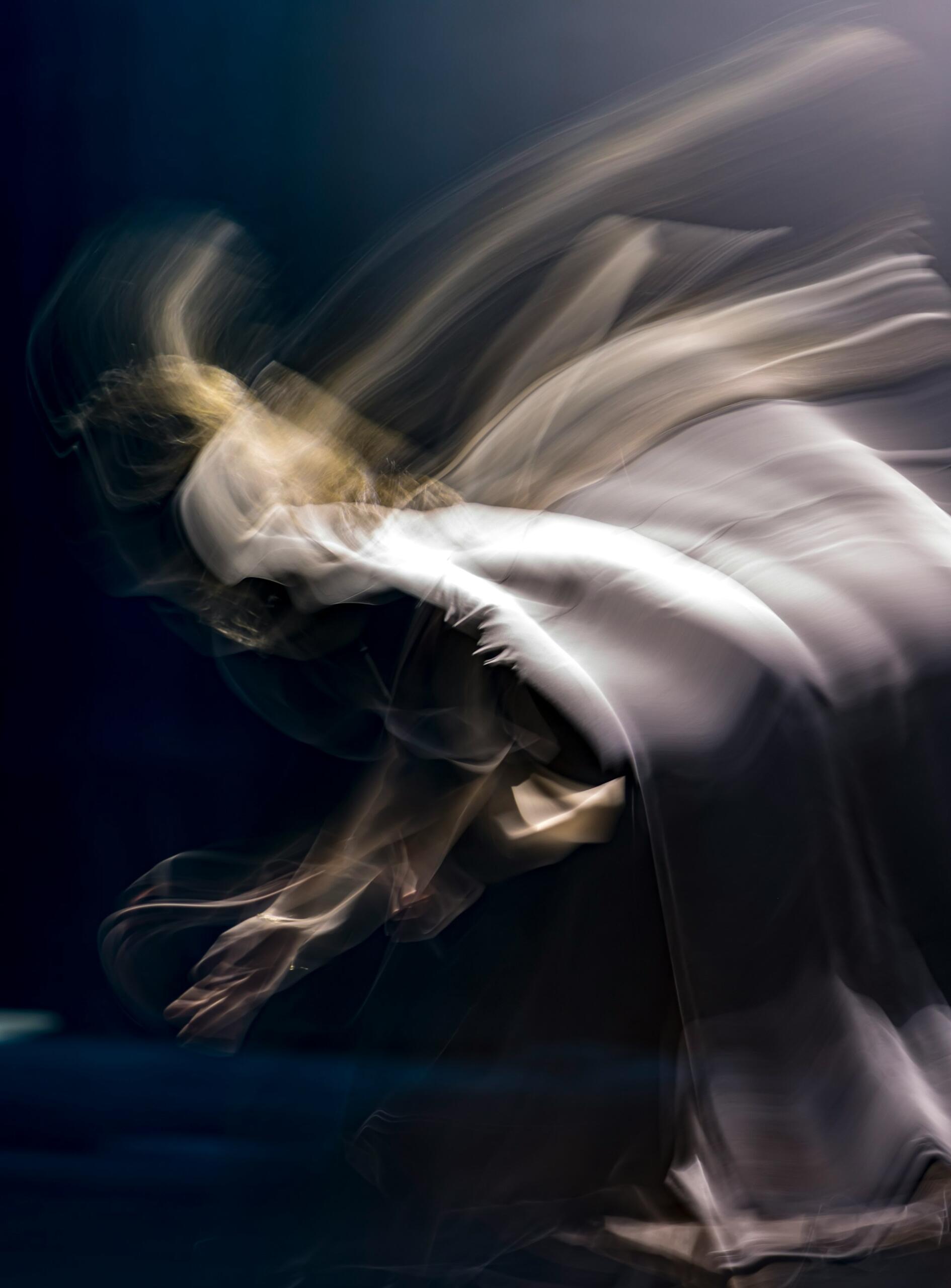 Una figura borrosa y fantasmal en movimiento, envuelta en una tela fluida, se mueve con elegancia sobre un fondo oscuro, evocando una sensación de movimiento etéreo.
