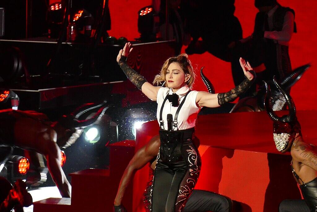Madonna con un traje de escenario blanco y negro con tirantes y guantes de encaje posa con los brazos levantados en un escenario iluminado con luz roja, rodeado de bailarines.
