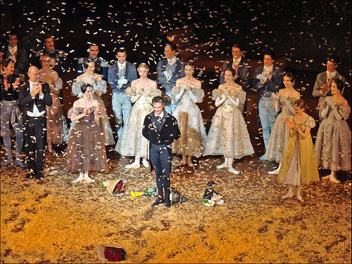 Manuel Legris se inclina en el escenario, rodeado por un elenco vestido con elegantes trajes de época, bajo una lluvia de confeti y con ramos de flores a sus pies, evocando una celebración y un triunfo.