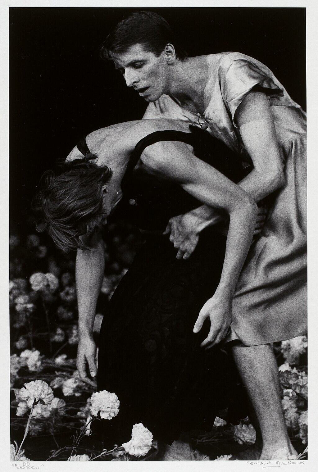 Dos bailarinas se abrazan emotivamente en un escenario cubierto de flores. Una, con un vestido oscuro, se inclina hacia delante, sostenida por la otra, con un vestido claro, transmitiendo dramatismo e intensidad.