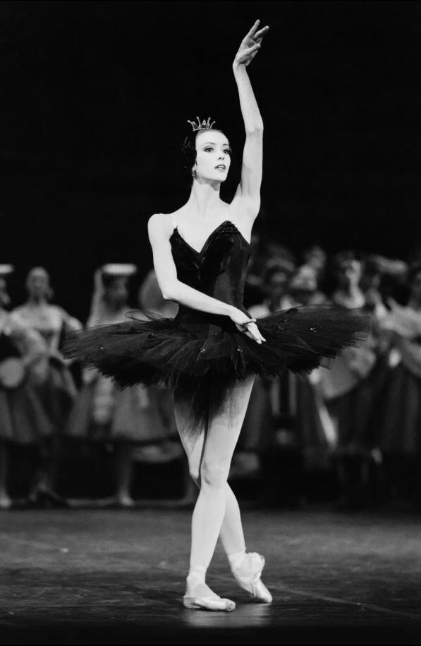 Sylvie Guillem con un tutú negro posa con elegancia en el escenario, con un brazo levantado en alto. Las figuras borrosas del fondo sugieren el escenario de una representación teatral.