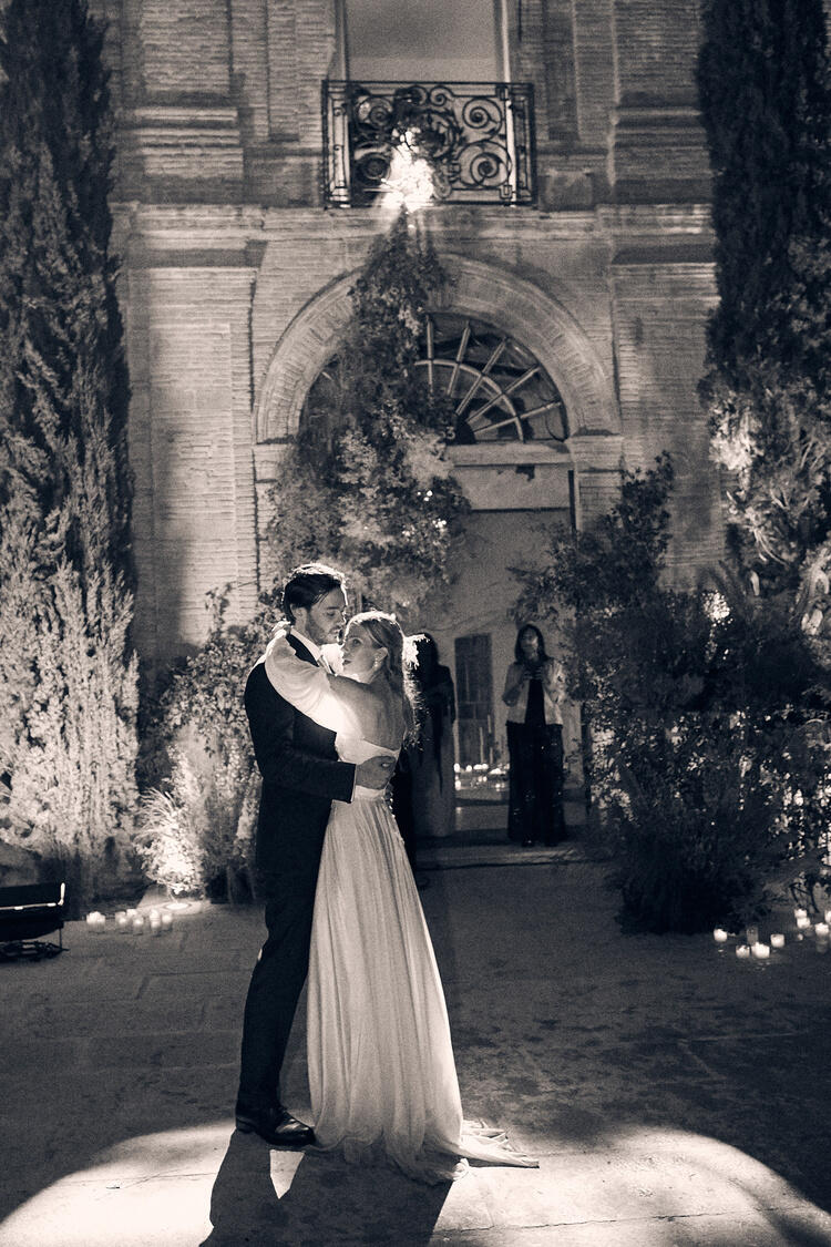 Una pareja vestida de novios baila románticamente bajo una suave iluminación, rodeada de vegetación y velas, con un elegante edificio de piedra como telón de fondo.