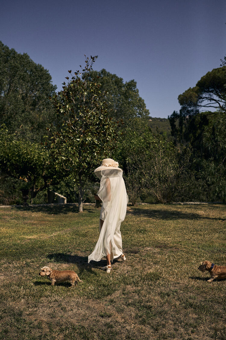 Una mujer con un vestido blanco vaporoso y un sombrero para el sol se aleja por un frondoso jardín acompañada de dos perritos. La escena transmite serenidad e idilio.