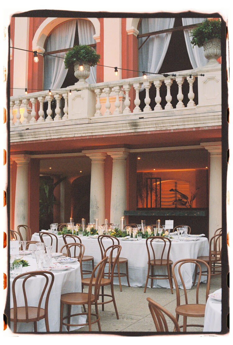 Elegante comedor al aire libre con mesas redondas, manteles blancos, sillas de madera y velas. El fondo muestra un balcón arqueado con plantas.