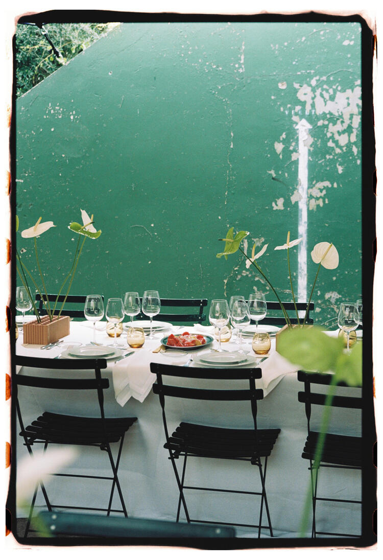 Elegante comedor al aire libre con sillas negras alrededor de una mesa adornada con manteles blancos, copas de vino y centros de mesa florales. El ambiente es acogedor y sereno.