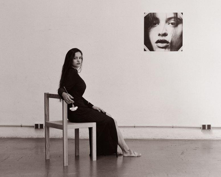 Rosalía con un vestido largo negro está sentada descalza en una silla de madera en una habitación minimalista. Sostiene una rosa blanca. En la pared hay un retrato en primer plano, lo que crea un ambiente contemplativo.