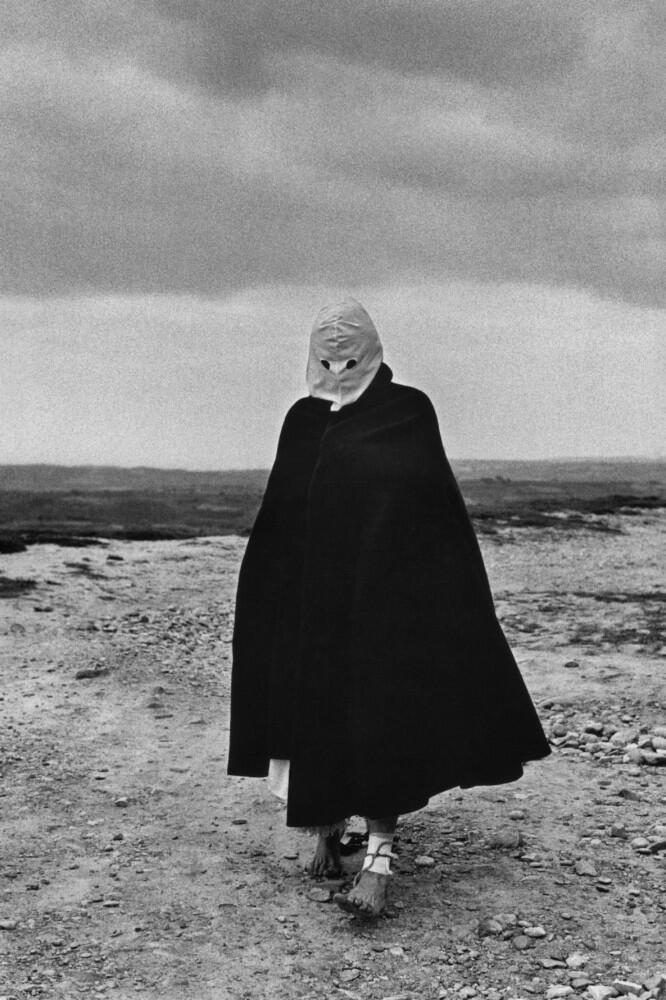 Una figura se encuentra de pie en un paisaje árido y rocoso, vestida con una capa oscura y una máscara blanca que le oculta el rostro. La escena resulta misteriosa bajo un cielo nublado.