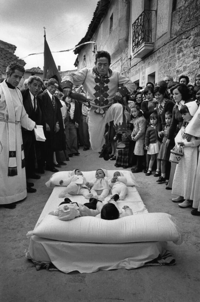 Un hombre con un traje festivo salta sobre un colchón con varios bebés, rodeado de curiosos en una calle estrecha. El ambiente es animado y tradicional.