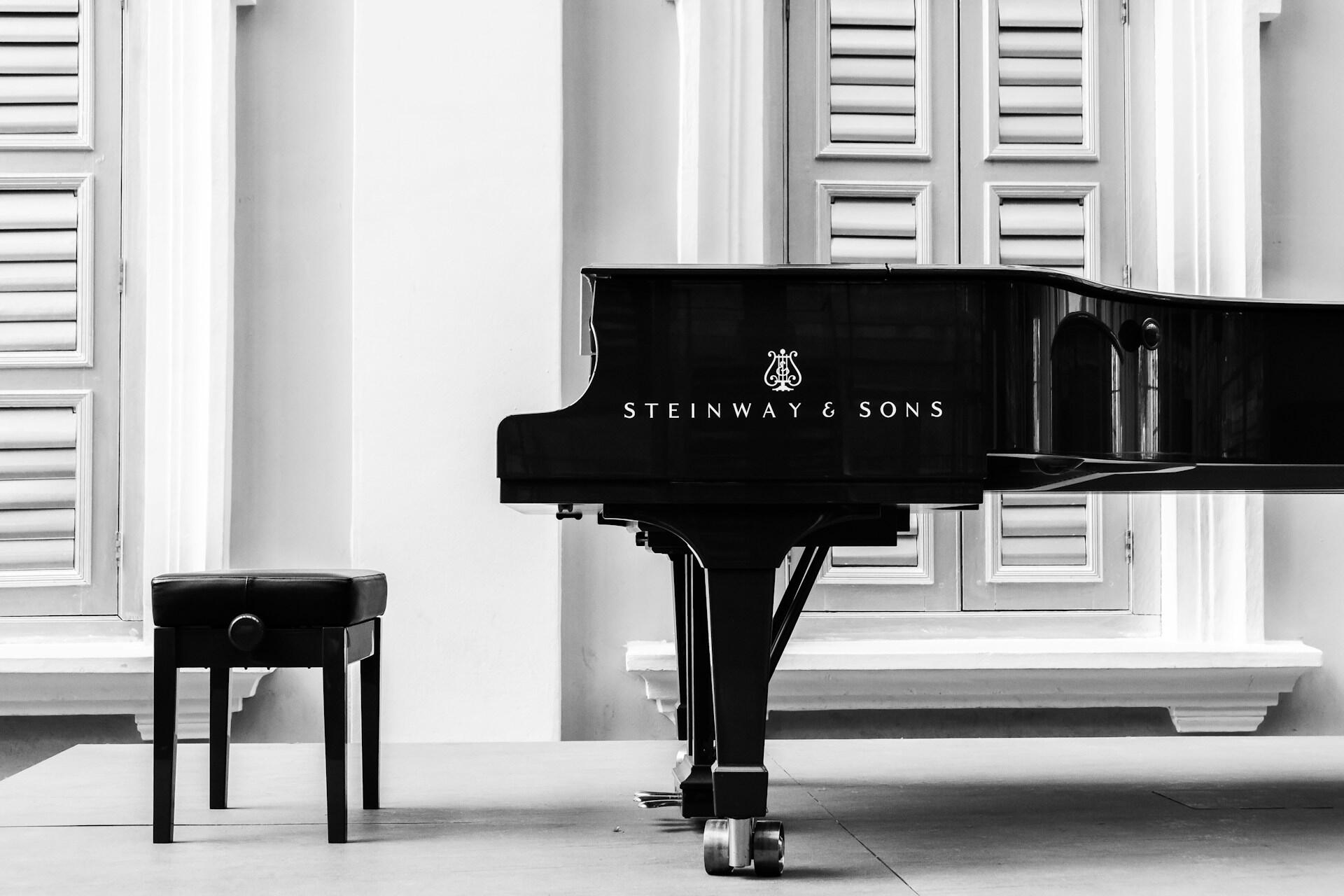 Imagen en blanco y negro de un piano de cola y un taburete con ventanas cerradas con contraventanas al fondo. El piano es un Steinway & Sons que irradia elegancia y sofisticación.