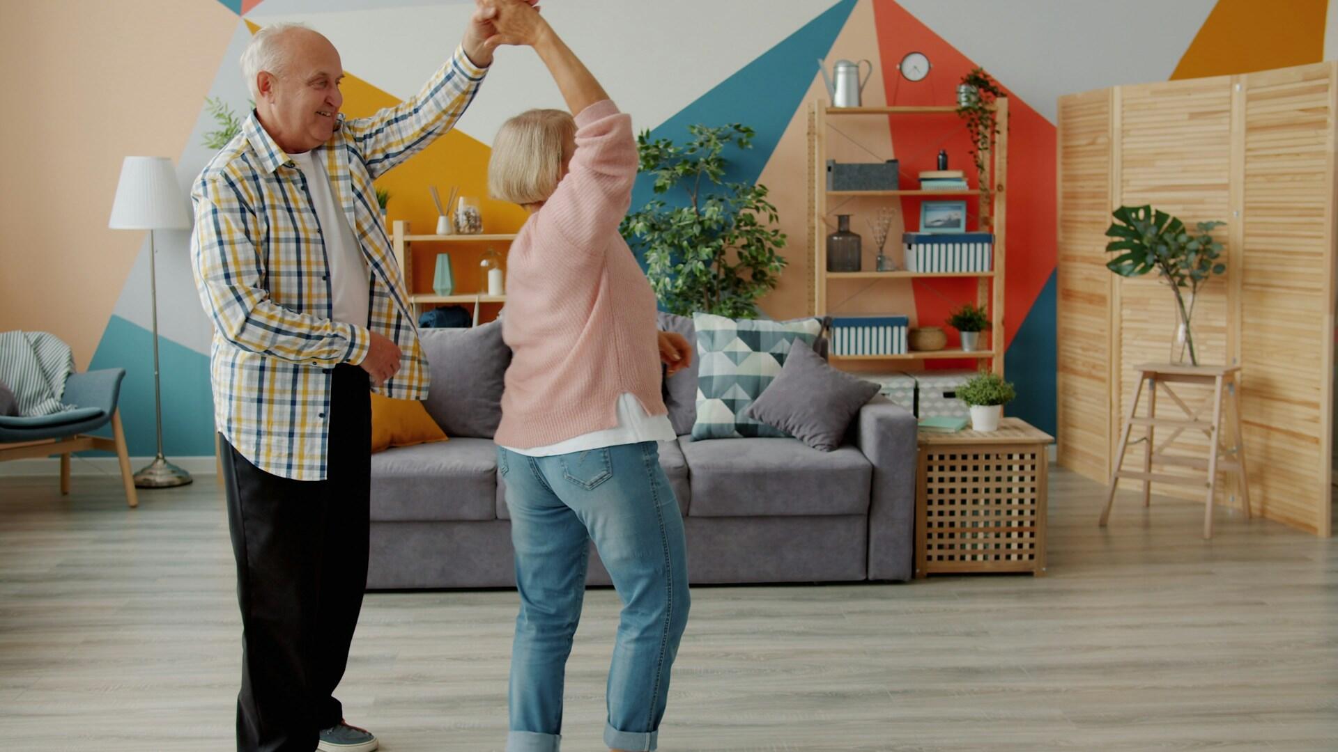 Pareja de ancianos bailando alegremente en una colorida sala de estar con motivos geométricos en las paredes, estanterías de madera y una decoración acogedora, que transmite calidez y felicidad.