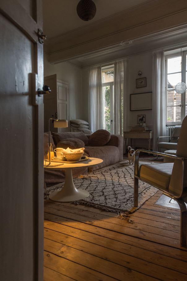 Una acogedora sala de estar con un sofá, sillas, una mesa de centro redonda con aperitivos, una alfombra estampada, suelos de madera y luz que entra por los grandes ventanales.