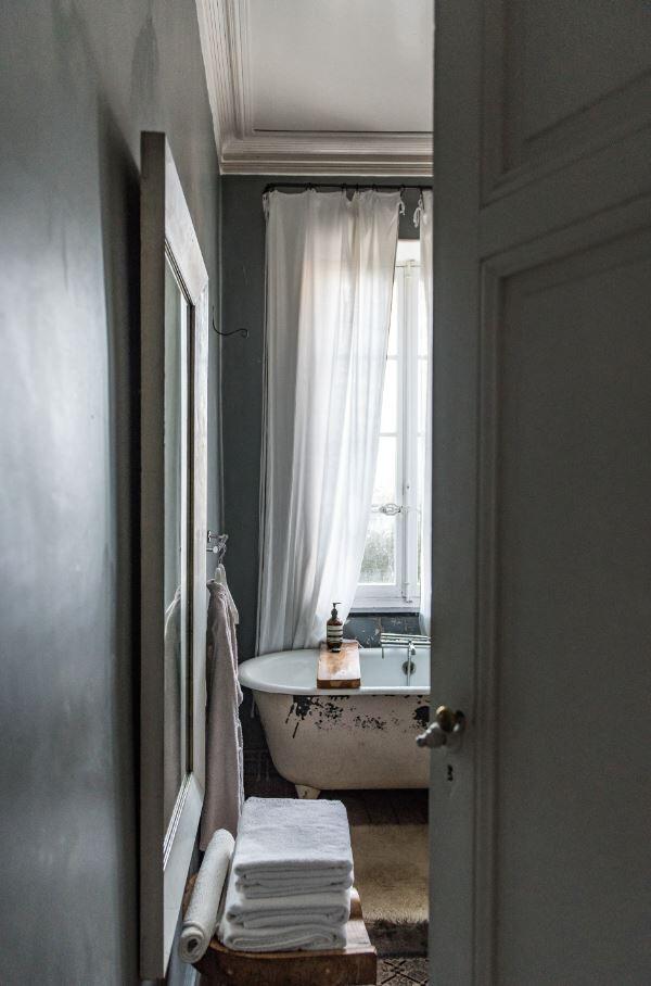 Un pequeño cuarto de baño vintage con una bañera con patas debajo de una ventana cubierta con cortinas blancas transparentes. Las toallas están cuidadosamente apiladas sobre un taburete, creando una sensación acogedora.