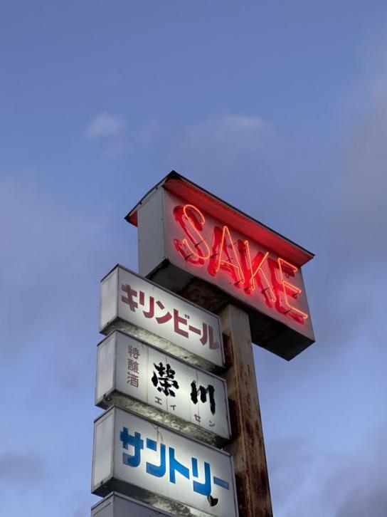 Al atardecer, contra un cielo nublado, se lee «SAKE» en neón rojo en un letrero alto. Debajo, se muestra texto en japonés en paneles iluminados, lo que transmite una atmósfera nostálgica.