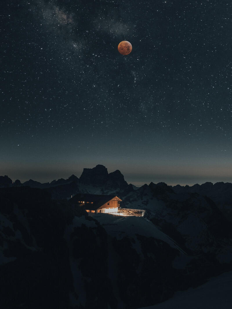 Una acogedora cabaña iluminada encaramada en una montaña se recorta contra un cielo nocturno estrellado, con una prominente luna rojiza, evocando tranquilidad y asombro.