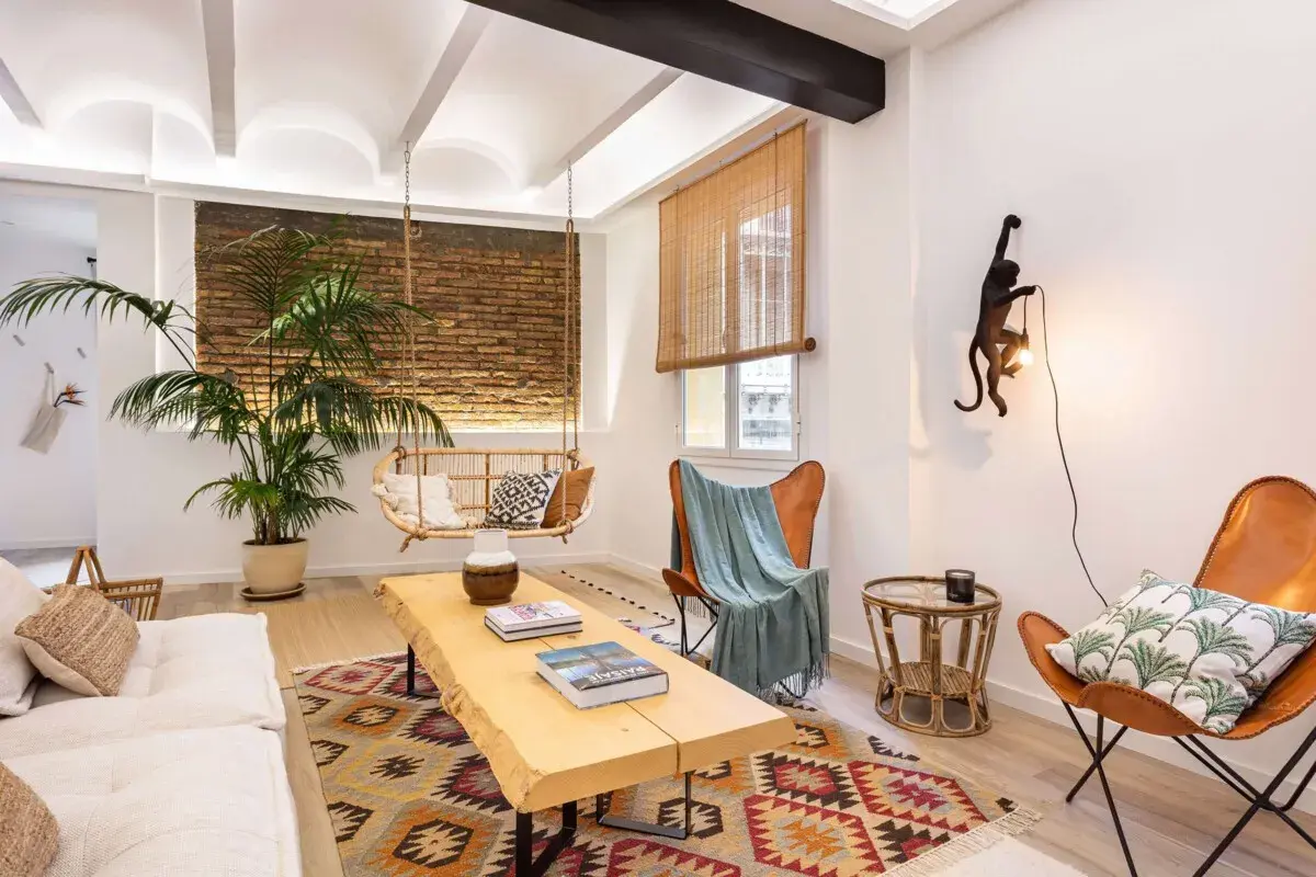 Acogedora sala de estar con un ambiente rústico, con una silla colgante, una mesa de centro de madera, sillas de cuero y una lámpara de pared con forma de mono. Tonos cálidos tierra.