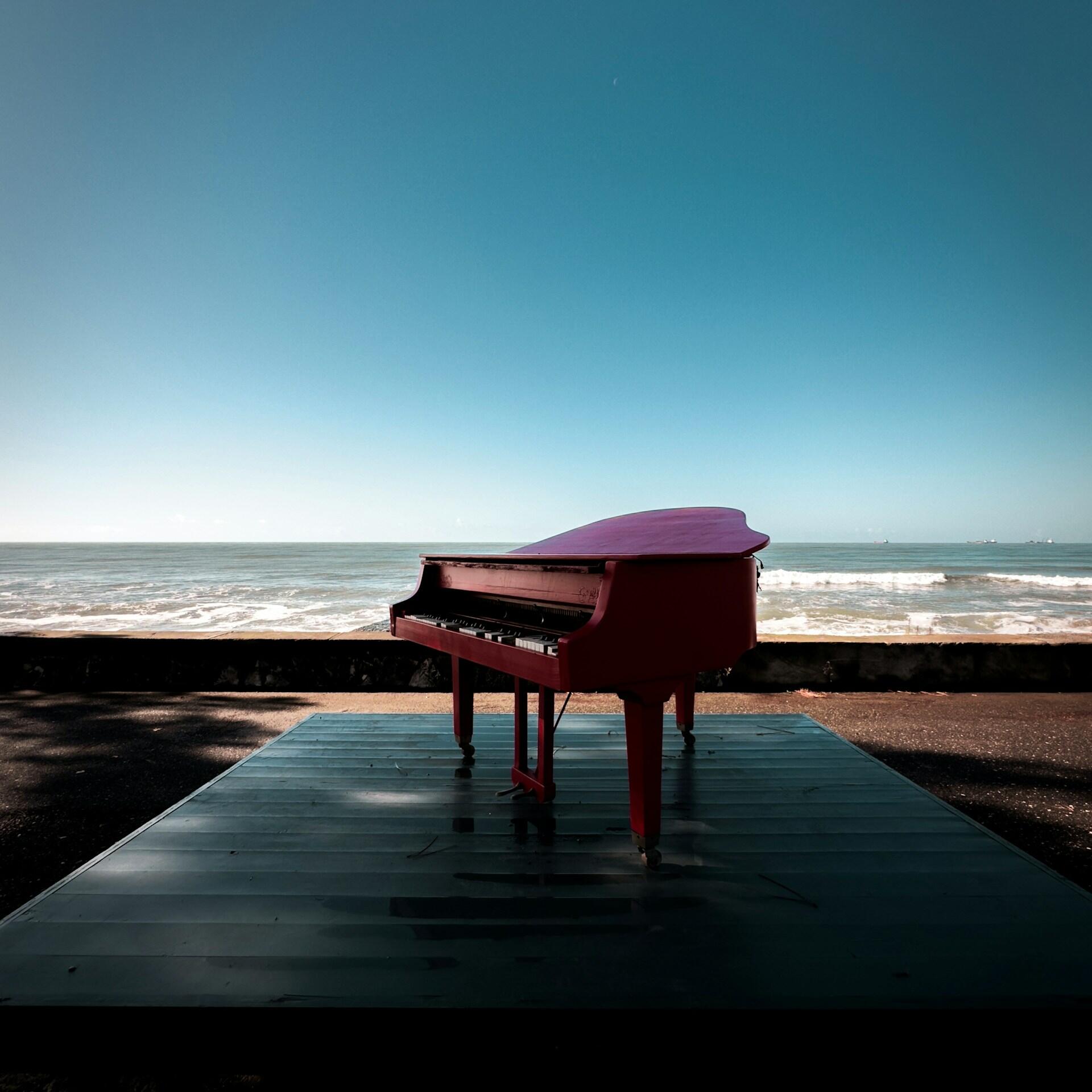 Un piano de cola rojo sobre una plataforma junto al mar en un día soleado. El inmenso cielo y las suaves olas crean una atmósfera serena y artística.