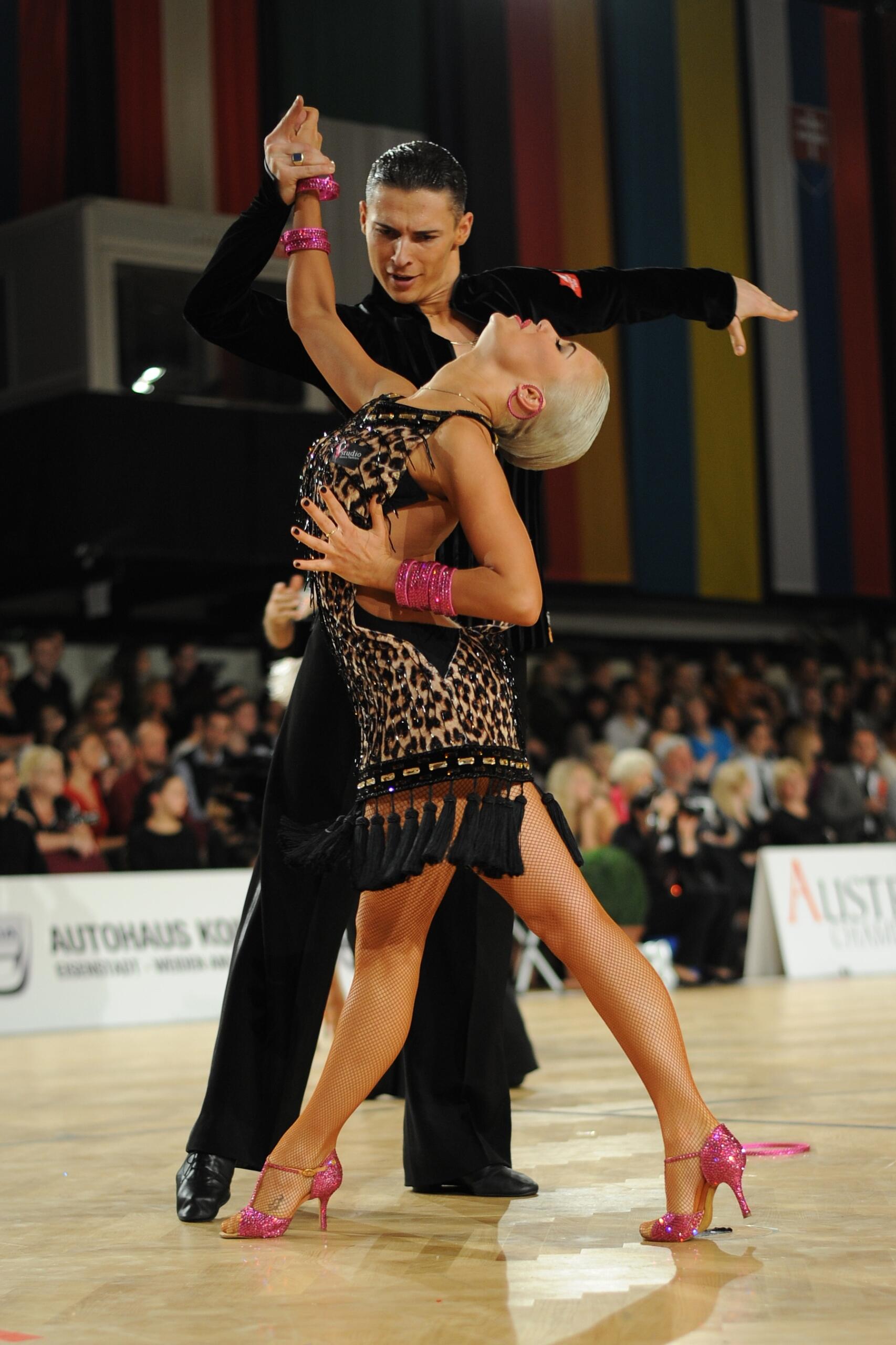 Los bailarines de salón bailan con pasión, con la mujer vestida con un vestido con estampado de leopardo y tacones rosas inclinándose hacia atrás sobre su pareja. El público, cautivado, observa.