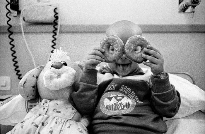 Un niño en una cama de hospital sostiene juguetonamente dos donas sobre sus ojos, sonriendo ampliamente, junto a un peluche de conejo. La imagen en blanco y negro transmite alegría e inocencia.
