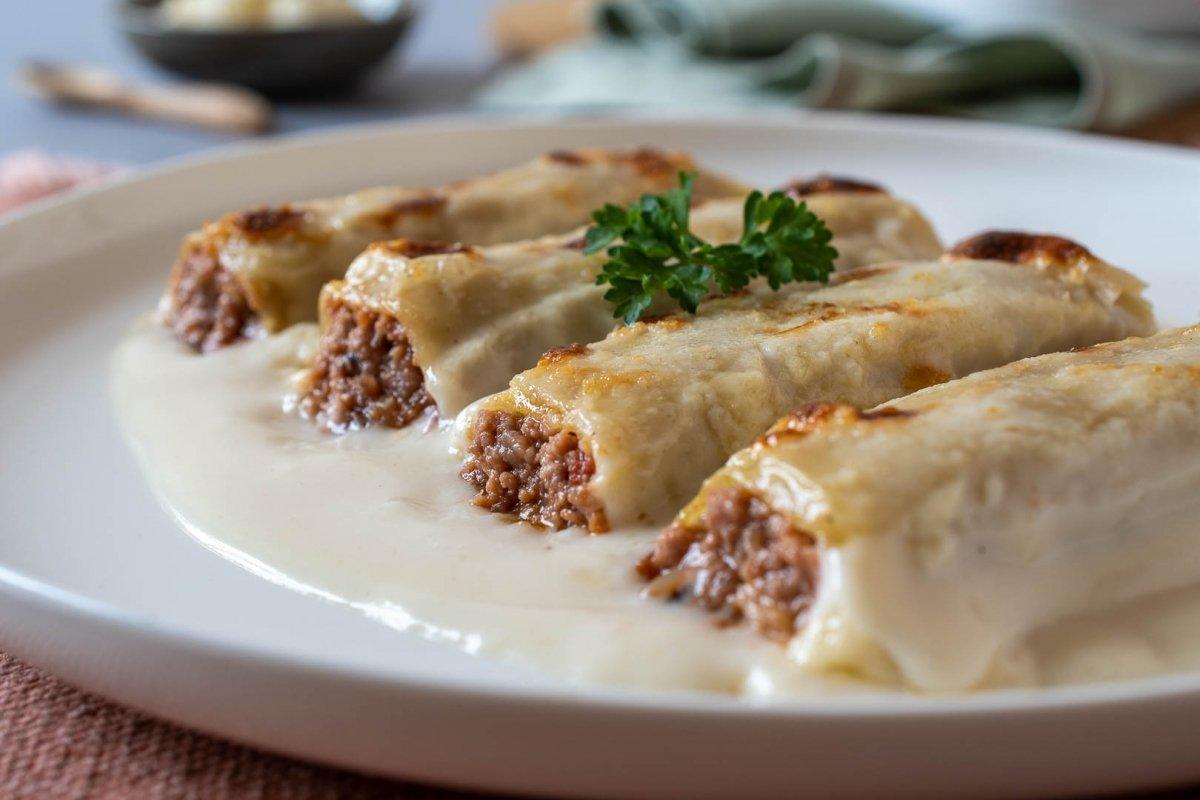 Canelones de carne servidos con abundante salsa blanca sobre plato blanco y decorados con un perejil.