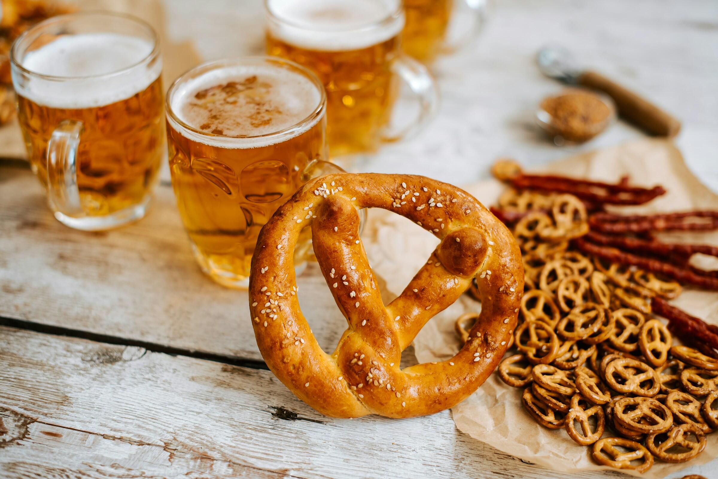 Un pretzel grande y suave con semillas de sésamo, rodeado de bocaditos y aperitivos de pretzel, con jarras de cerveza al fondo.