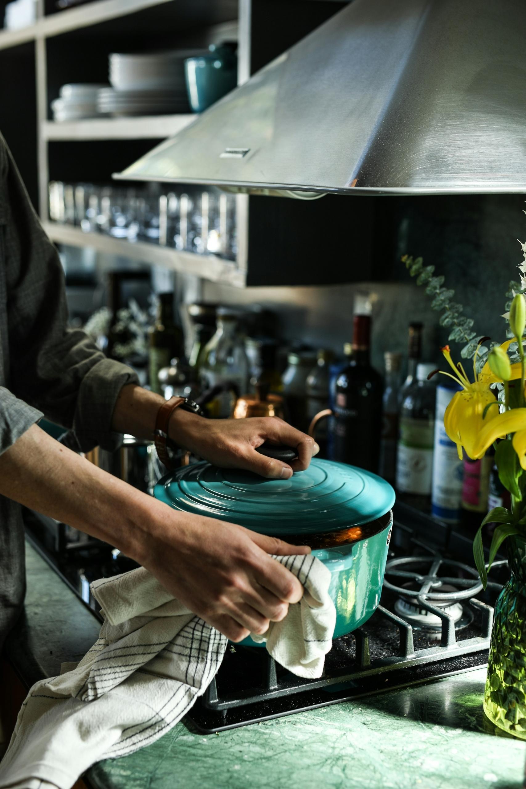 Persona sujetando una tapa de olla verde azulado en una cocina con estanterías y botellas al fondo.