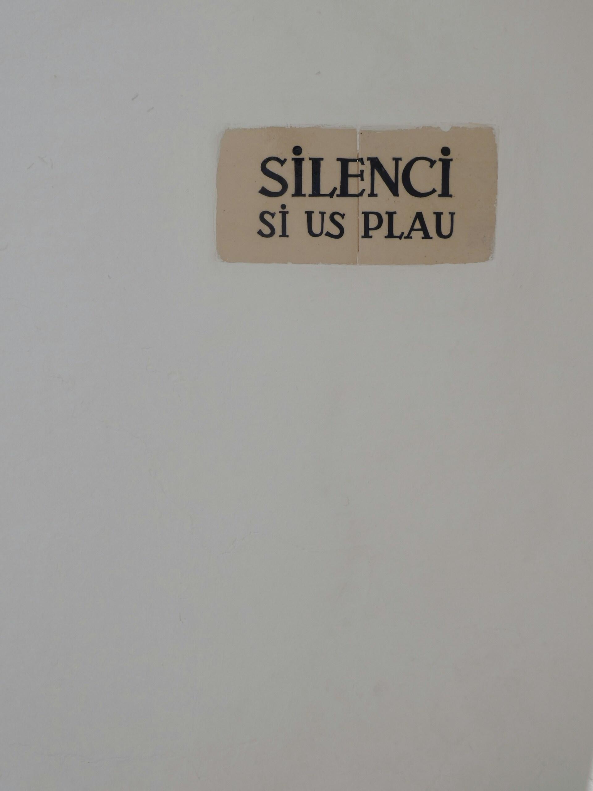Un pequeño cartel beige en una pared blanca lisa reza «SILENCI SI US PLAU» en letras mayúsculas negras, transmitiendo una cortés petición de silencio escrita originalmente en catalán.