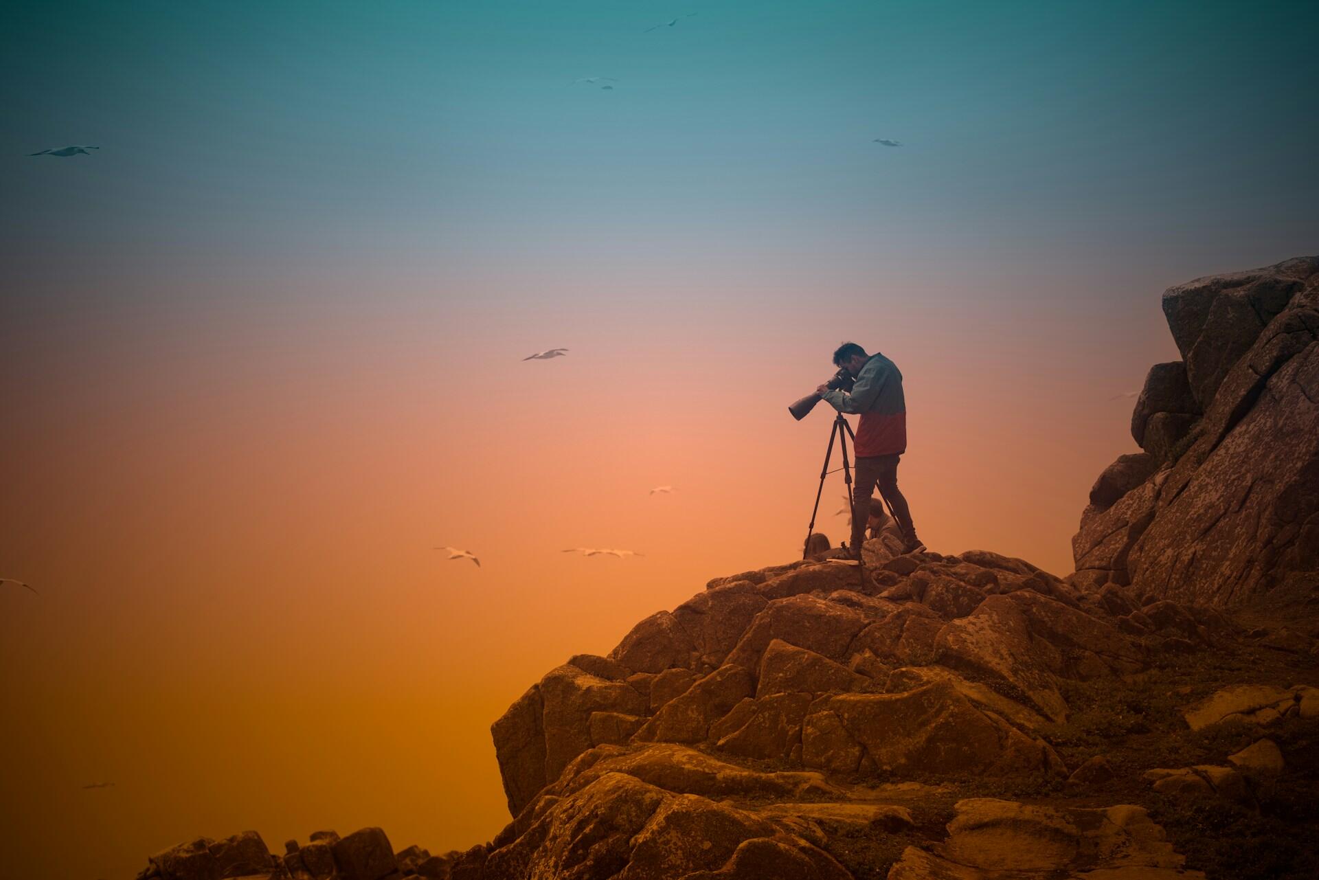 Un fotógrafo, en un terreno rocoso, utiliza una cámara montada en un trípode con el cielo de una puesta de sol de colores vivos como telón de fondo. Las gaviotas vuelan al fondo, evocando tranquilidad y concentración.