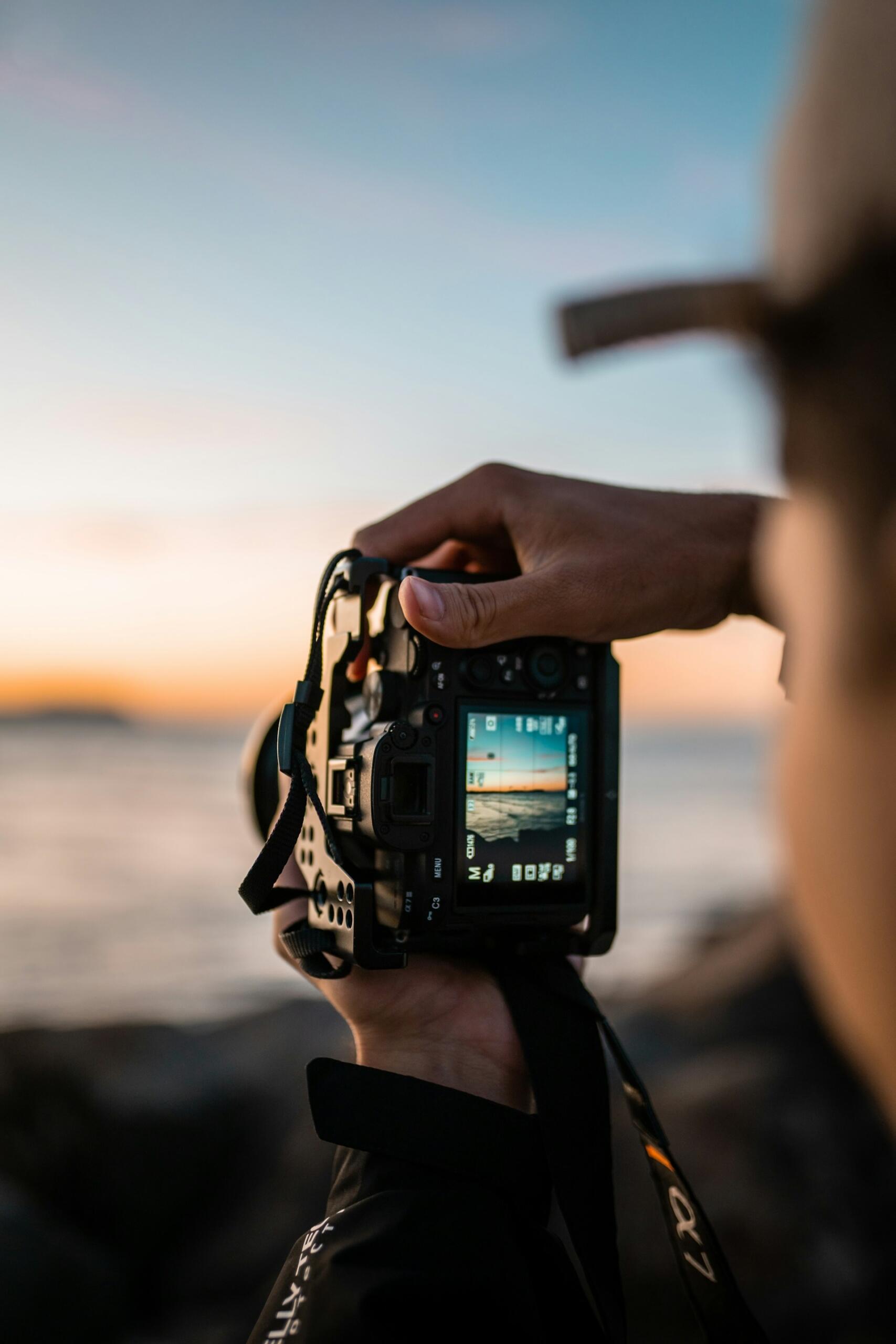 Las manos de un fotógrafo sostienen una cámara enfocada hacia una puesta de sol sobre el océano; en la pantalla de la cámara se aprecia la pintoresca vista. El ambiente es sereno y contemplativo.