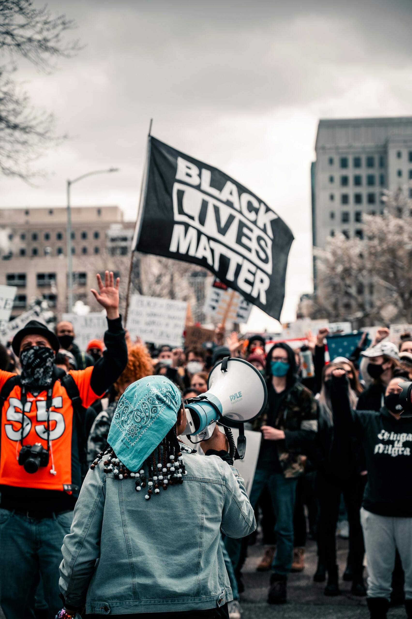 Un manifestante con un pañuelo azul habla por un megáfono frente a una multitud que enarbolaba una bandera con el lema «Black Lives Matter». El ambiente es apasionado y de unidad.