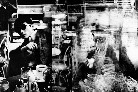 Fotografía en blanco y negro de una persona en una cafetería, borrosa y misteriosa. Los reflejos en el cristal aportan complejidad y crean una atmósfera reflexiva y artística.