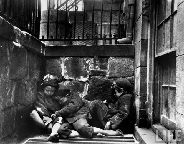 Tres niños se apiñan en un rincón de un callejón de piedra, con aspecto cansado y angustiado. La escena transmite una atmósfera urbana sombría y cruda.