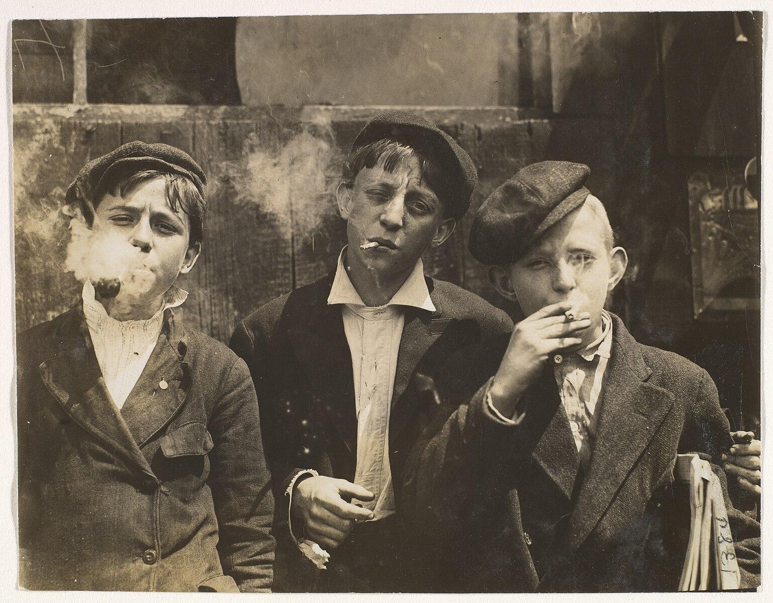 Tres chicos, con gorras planas y trajes, están de pie muy juntos, fumando cigarrillos. El tono sepia le da a la imagen un aire histórico y crudo.