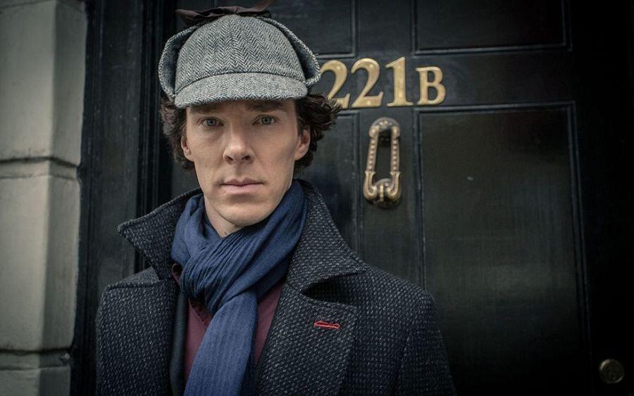 Benedict con un sombrero y un abrigo oscuro se encuentra de pie, solemne, frente a una puerta con el número 221B, evocando un ambiente misterioso y reflexivo.