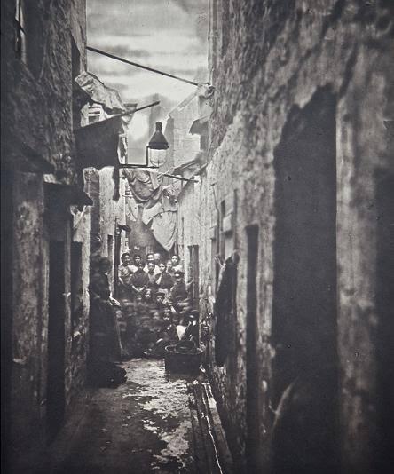 Un grupo de personas se reúne en un callejón estrecho y poco iluminado, flanqueado por edificios de piedra. Hay ropa colgada por encima de sus cabezas y se respira un ambiente sombrío y histórico.