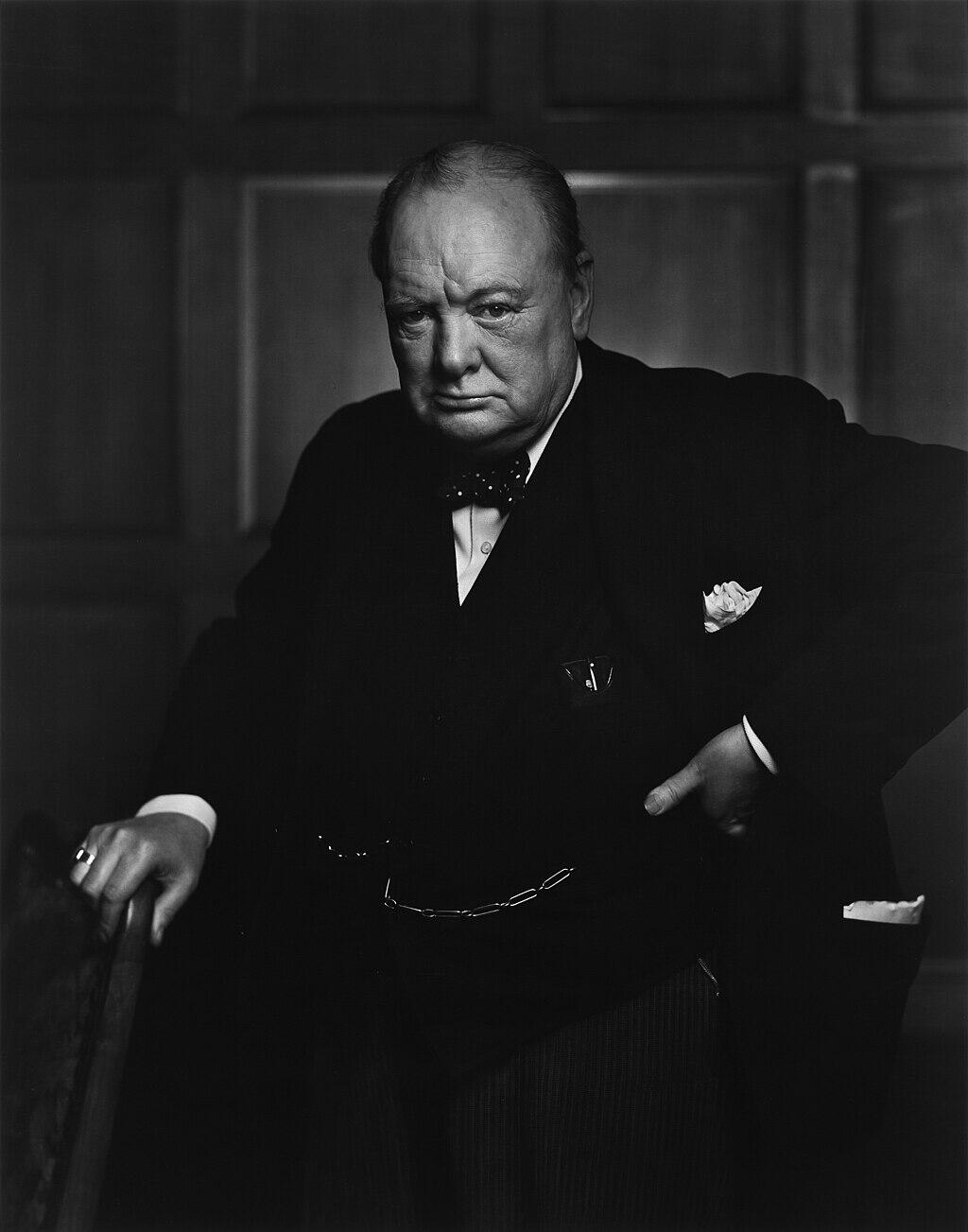 Un Winston Churchill de aspecto severo vestido con traje posa frente a un panel de madera. Sostiene una silla y apoya una mano en la cadera, irradiando confianza y autoridad.