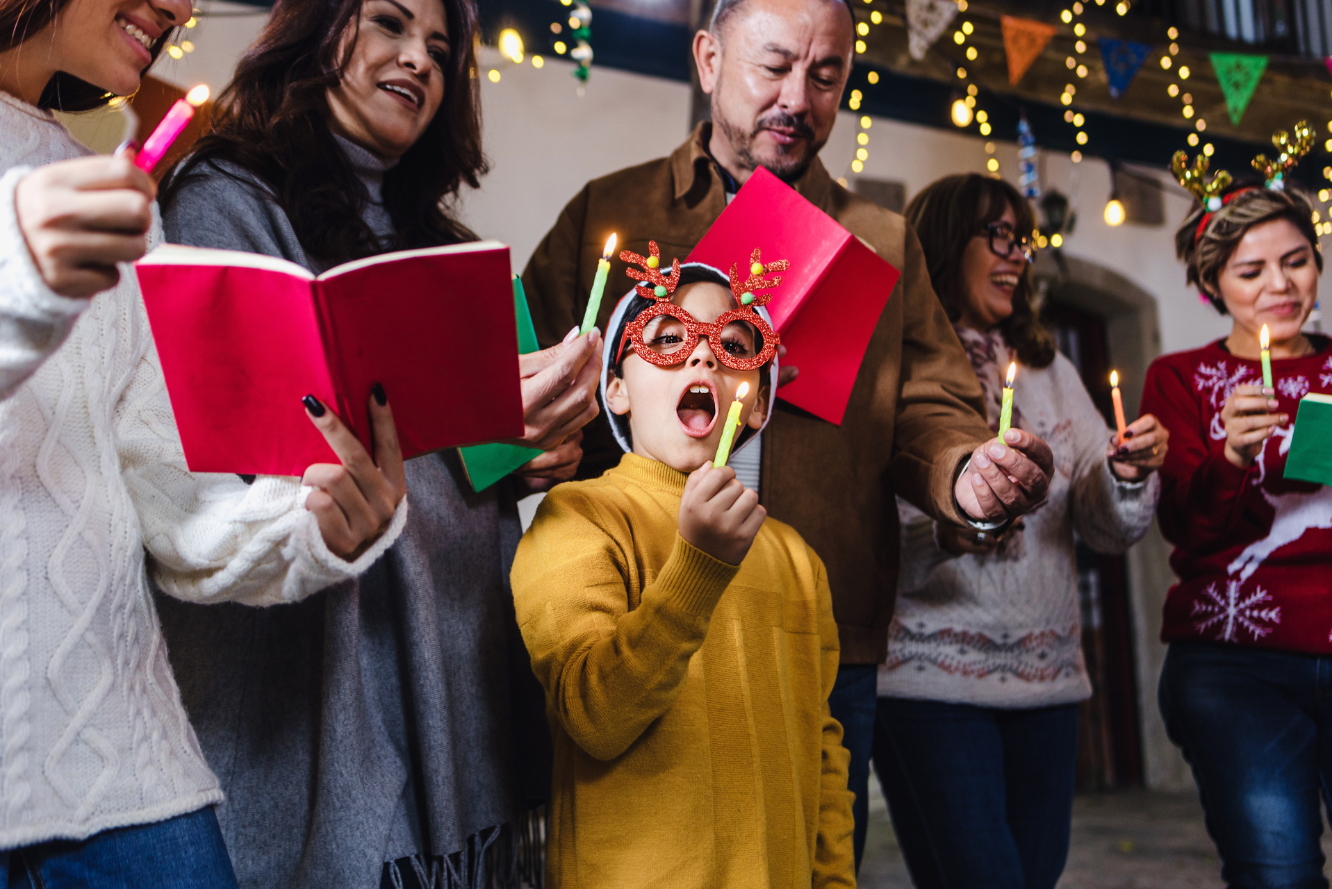 Grupo de personas sosteniendo velas y cancioneros, atuendo festivo, con luces navideñas de fondo.