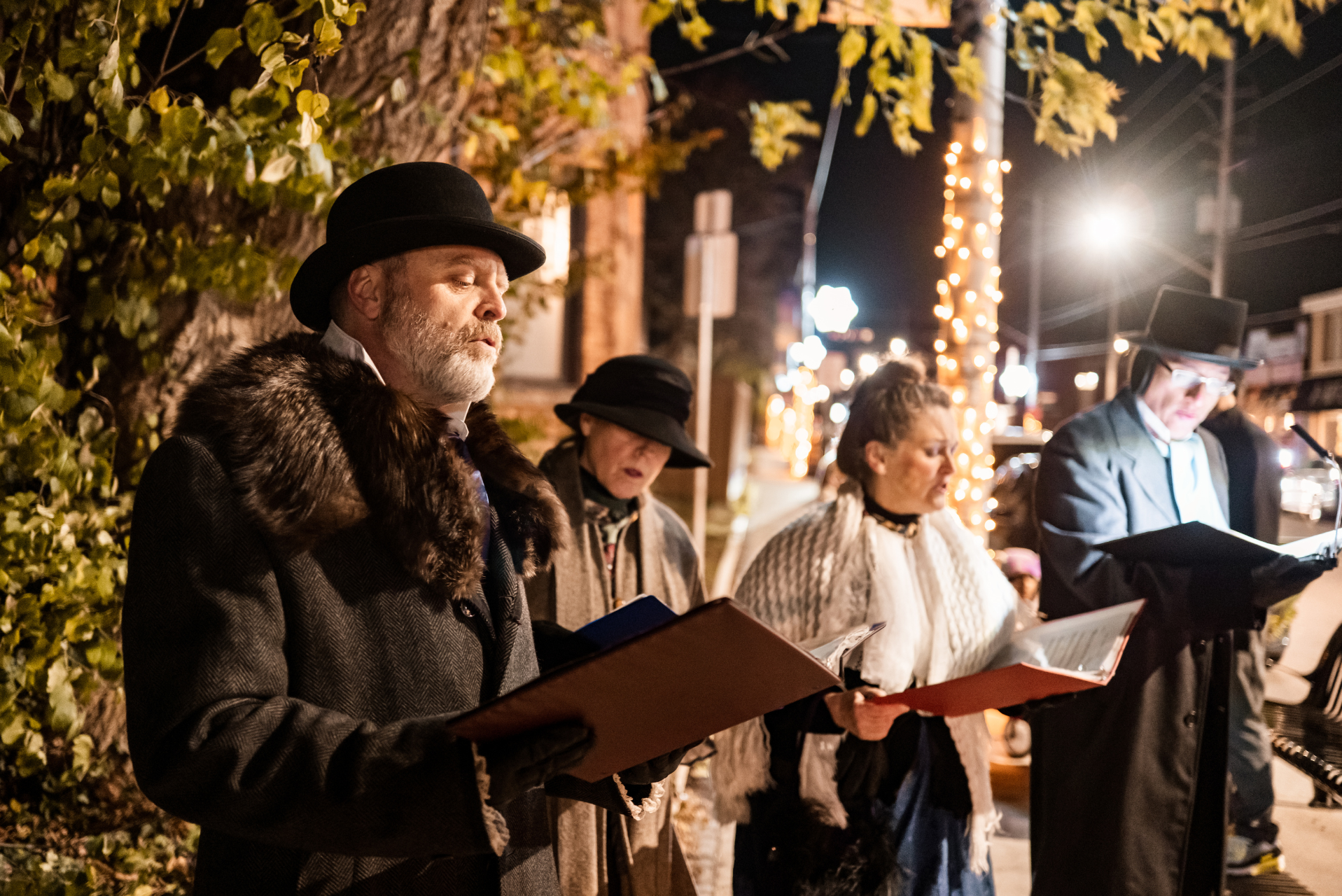 Un grupo de cantores de villancicos con cancioneros en la mano, de noche, con farolas y adornos de fondo.