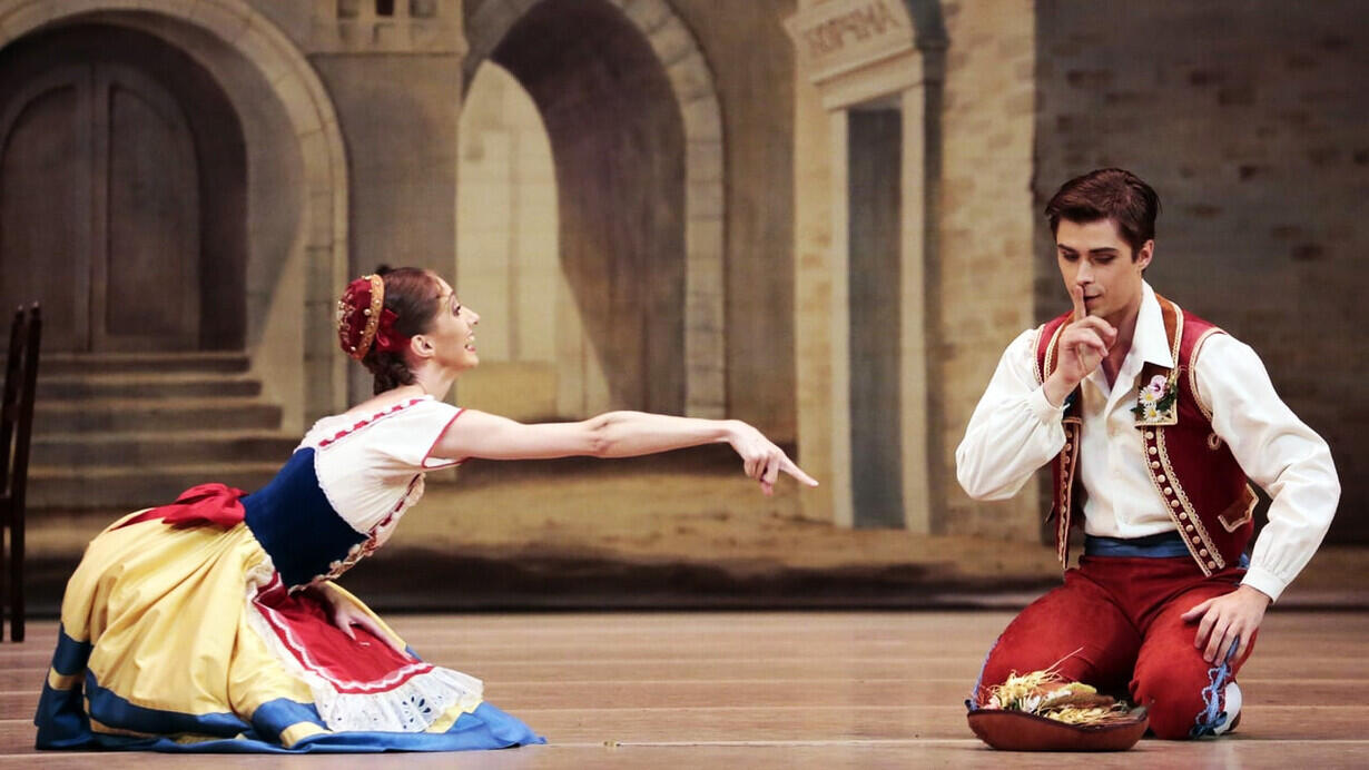 Una bailarina y un bailarín, vestidos con trajes tradicionales, actúan en el escenario. La mujer gesticula con entusiasmo mientras el hombre se arrodilla, creando un ambiente alegre.