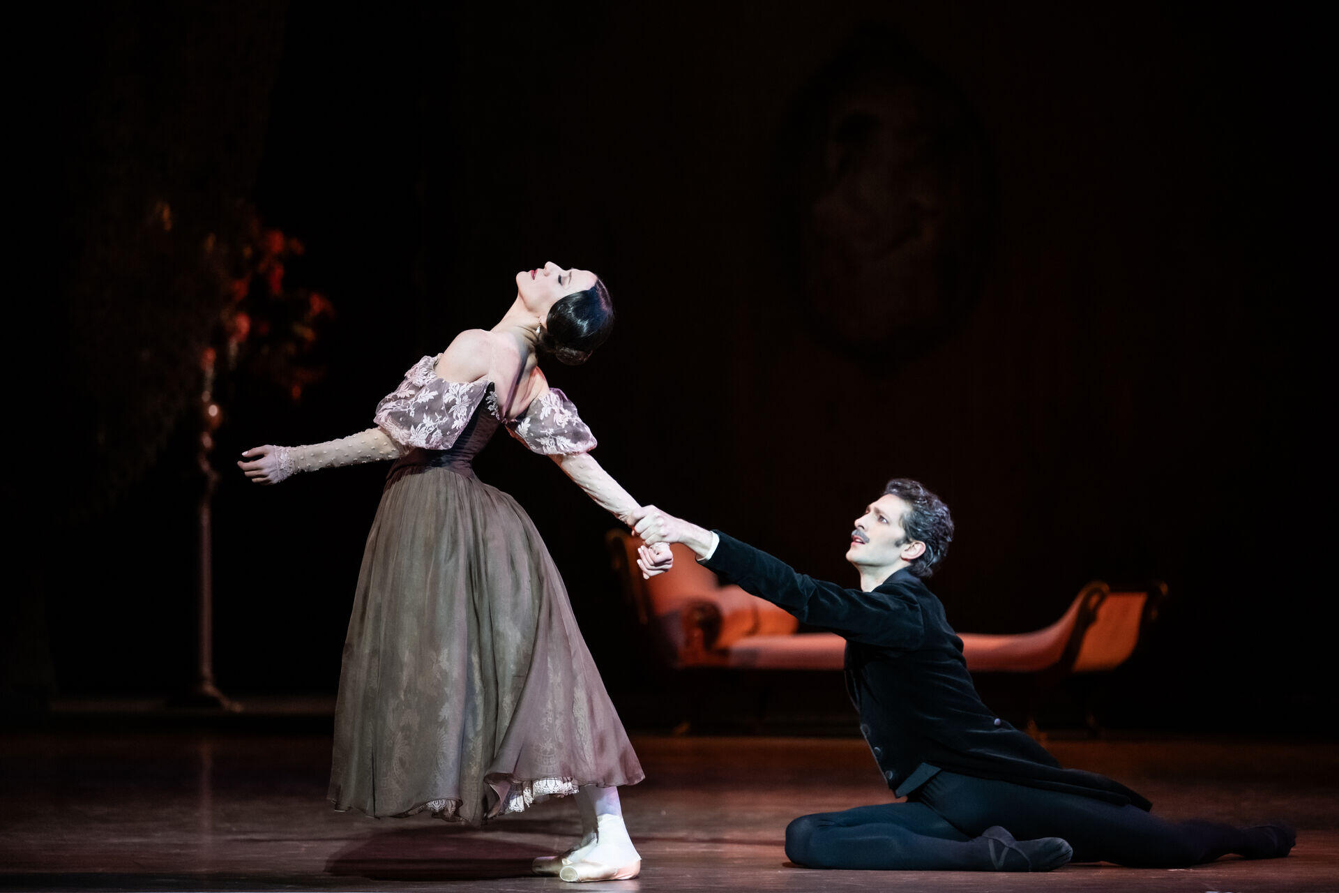 Una bailarina de ballet con un vestido vaporoso se inclina hacia atrás con elegancia mientras otro bailarín, vestido con un traje oscuro, se arrodilla y le toma la mano. La escena transmite elegancia y dramatismo.