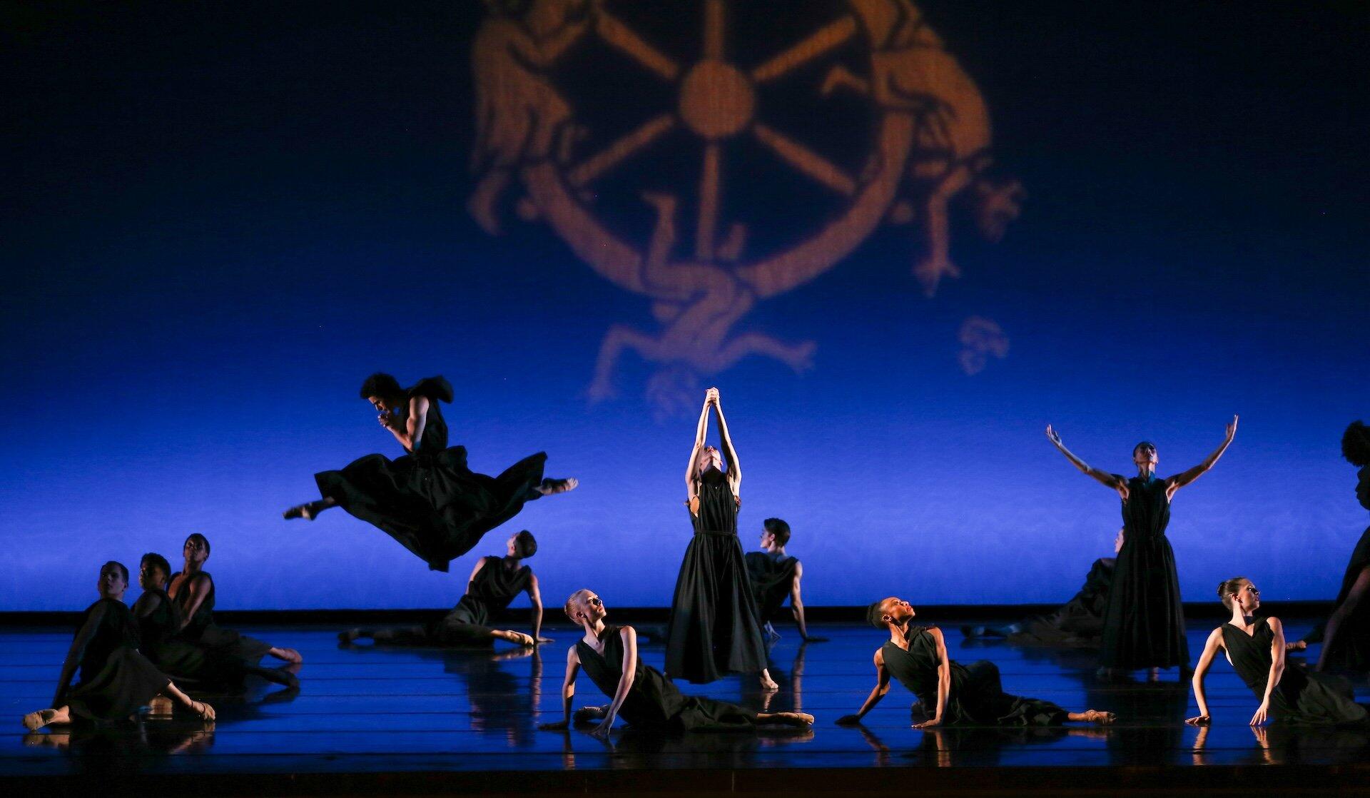 Un grupo de bailarinas vestidas de negro actúa con gran expresividad sobre el escenario, y una de ellas salta en el aire. Sobre un fondo azul se proyecta el símbolo de una rueda dorada.
