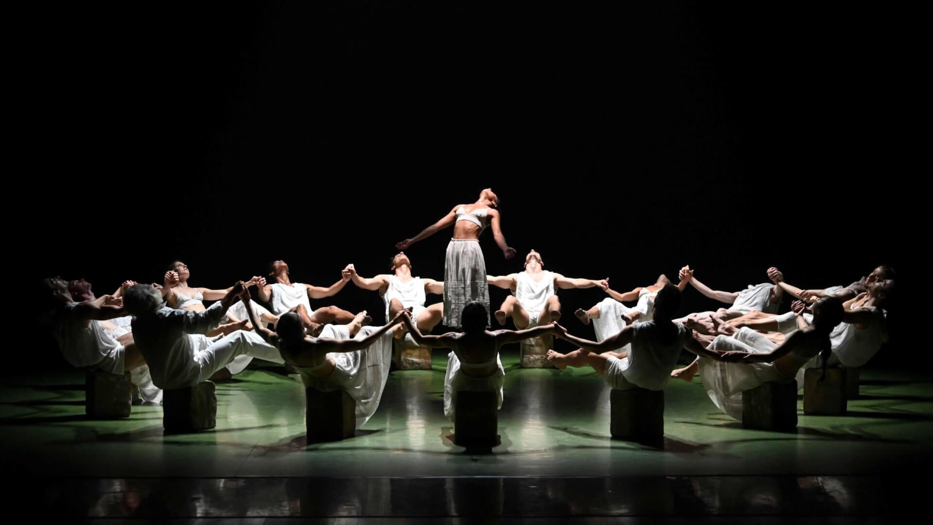 Un grupo de bailarines vestidos de blanco yacen boca arriba, formando un círculo sobre el escenario. Un bailarín se encuentra de pie en el centro, con los brazos extendidos, irradiando elegancia.